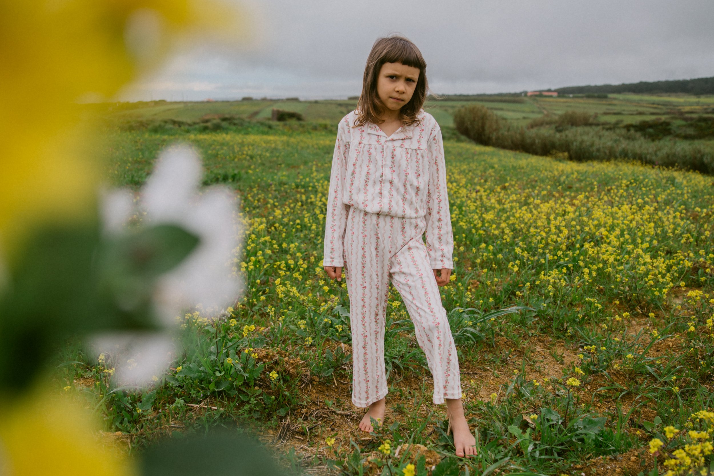 A young girl in pajamas stands barefoot in a field of yellow flowers, looking at the camera with a serious expression, under a cloudy sky.