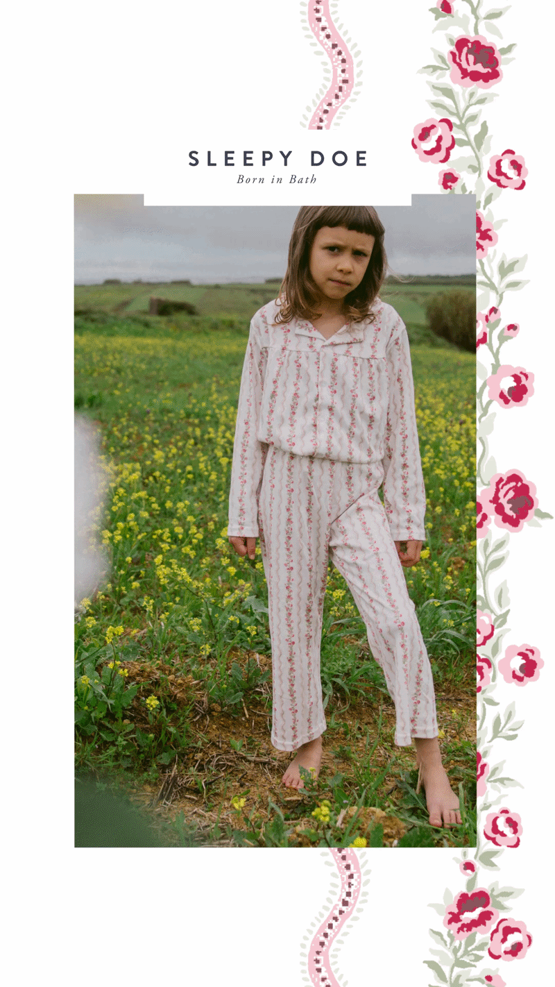 A young girl standing barefoot in a field of yellow flowers, wearing pink patterned pajamas with a serious expression.