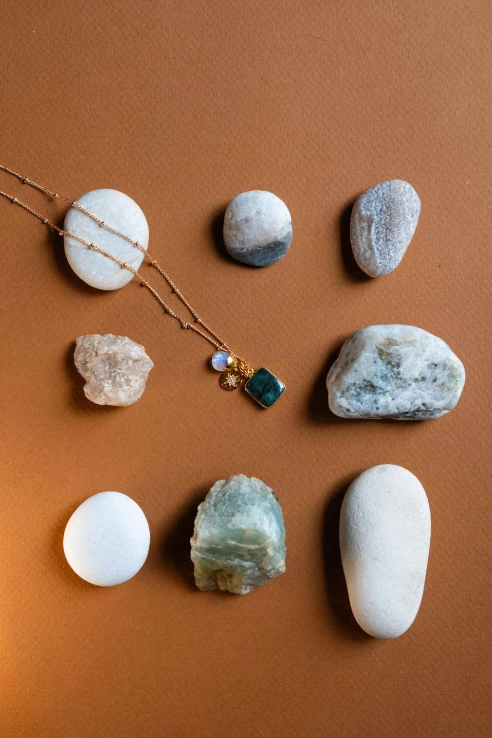 A collection of various stones and rocks with a gold necklace featuring a green square pendant and charms, arranged on a brown surface.