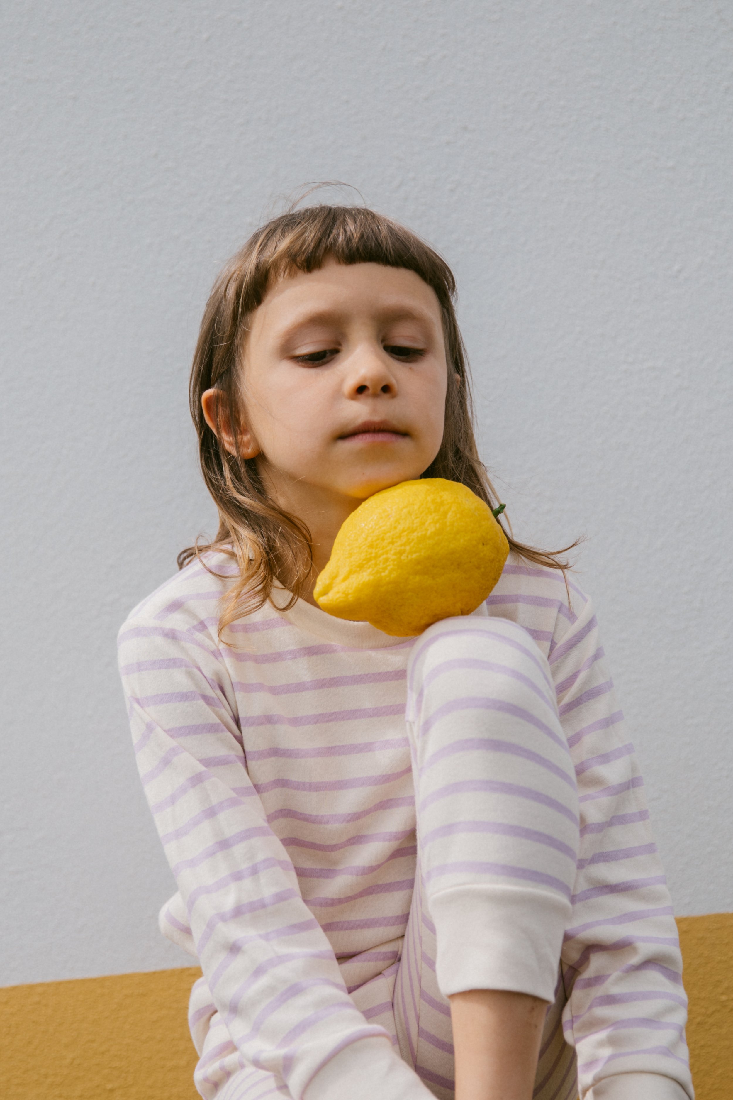 A young girl with light brown hair and bangs is holding a large yellow lemon in front of her face, looking down at it, against a background with a white wall and beige carpet.