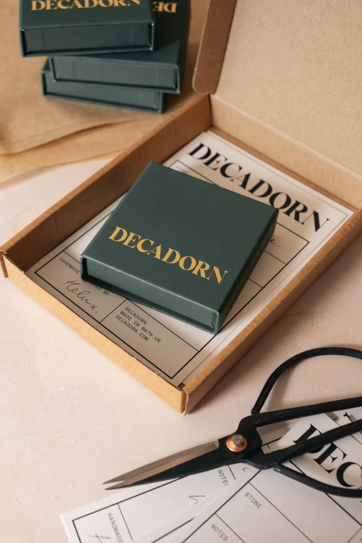 Open cardboard box with a small green box labeled 'Decadorn' inside, along with printed sheets of paper and scissors nearby. The green box also has 'Decadorn' written on it in gold lettering.
