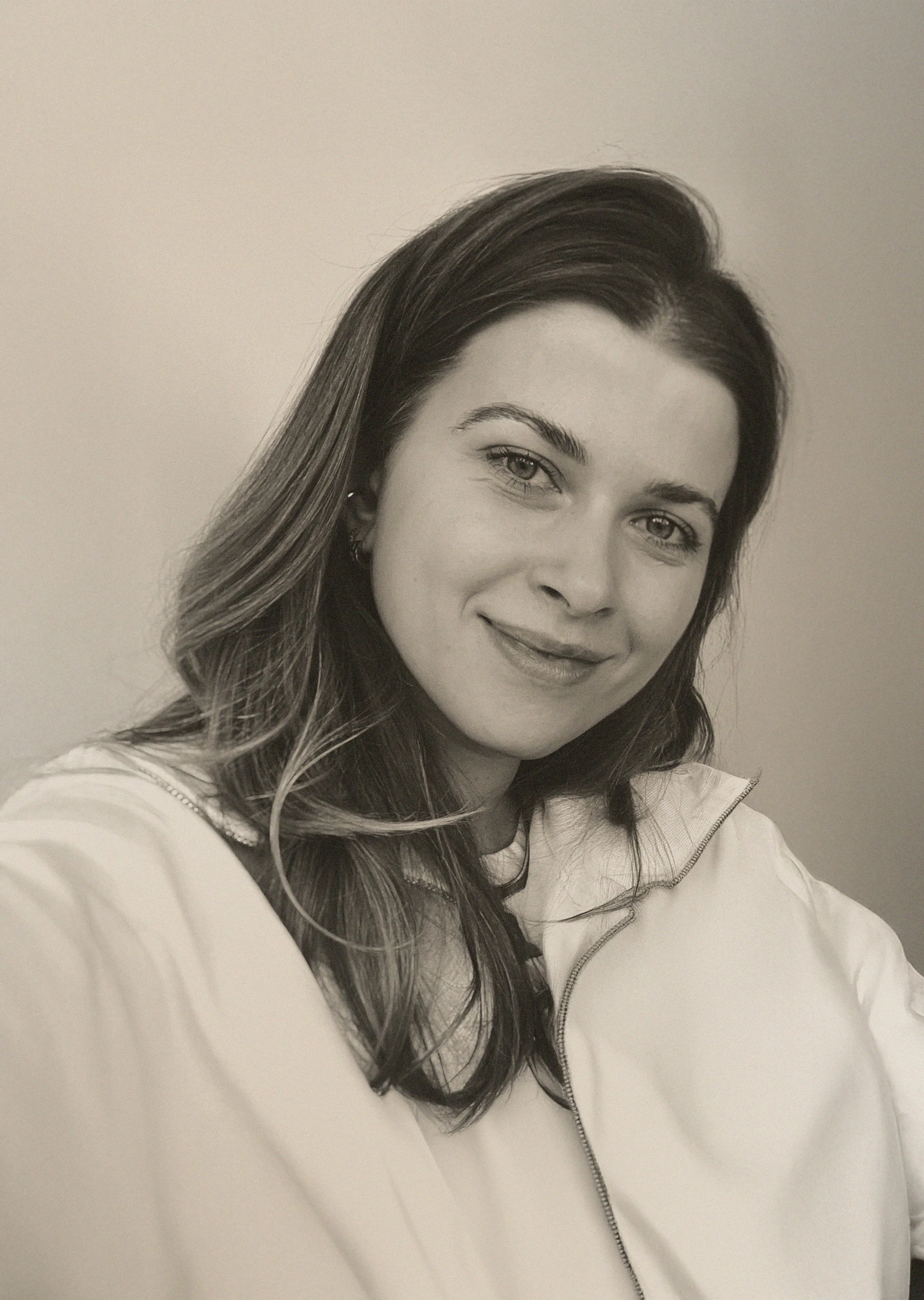 A woman with dark wavy hair, light eyes, and a friendly smile taking a selfie against a plain, light-colored wall. She is wearing a light-colored jacket with a zipper.