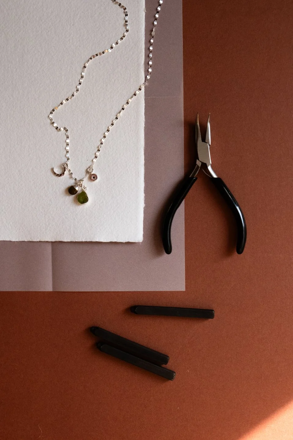 Silver necklace with four small, colorful charms and a moon and star design, on white foam, with jewelry pliers and black jewelry clips on a reddish-brown surface.