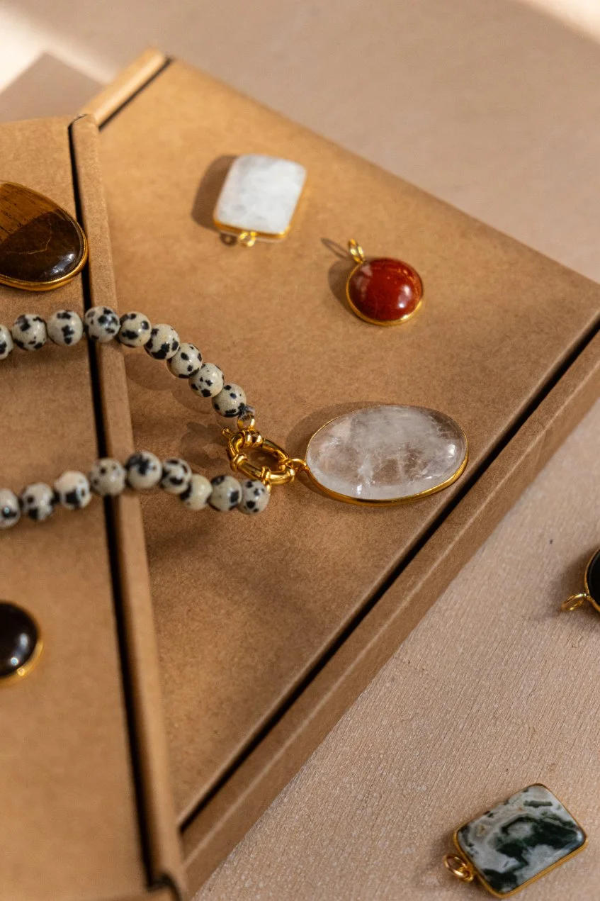 Various jewelry pieces, including a beaded bracelet, a pendant with a clear stone, a red gemstone pendant, and a white rectangular pendant, displayed on a cardboard jewelry tray.