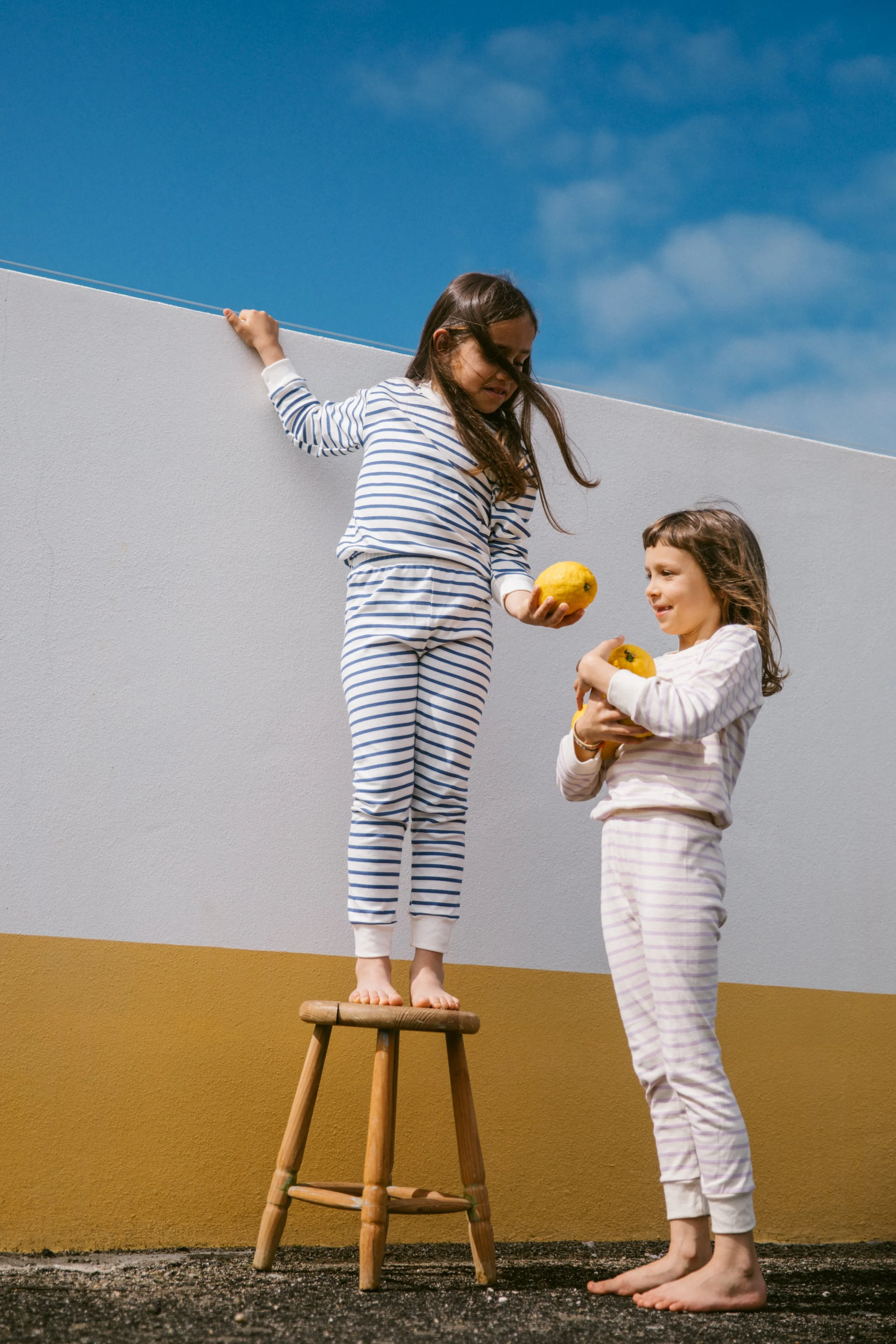 Two young girls in striped pajamas balancing on a stool outdoors, holding yellow fruit, with a yellow and white wall and blue sky behind them.