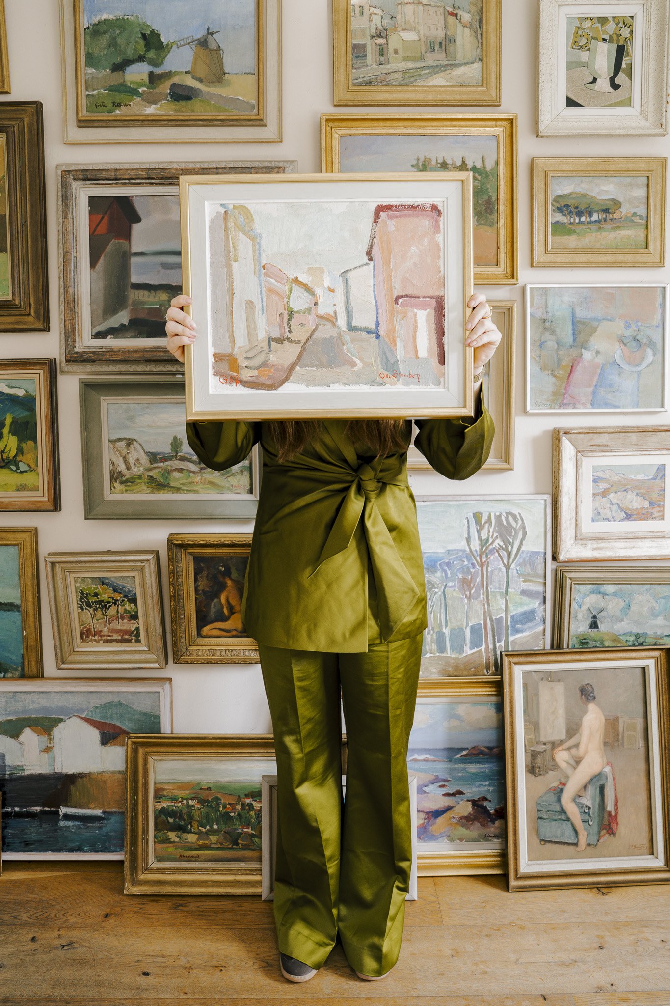 Person holding a framed painting in front of a wall filled with various framed artworks in an art gallery or studio.