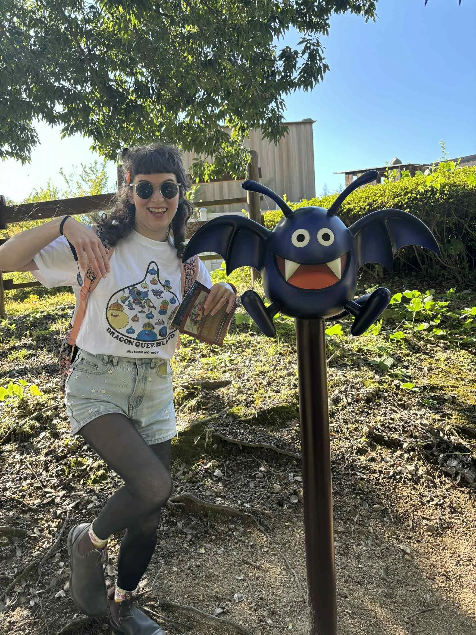 A woman wearing sunglasses, a white 'Dragon Quest Island' t-shirt, shorts, and black tights is standing outdoors next to a dragon-shaped sign with big eyes, wings, and an open mouth, in a garden area with trees and a wooden building in the background.
