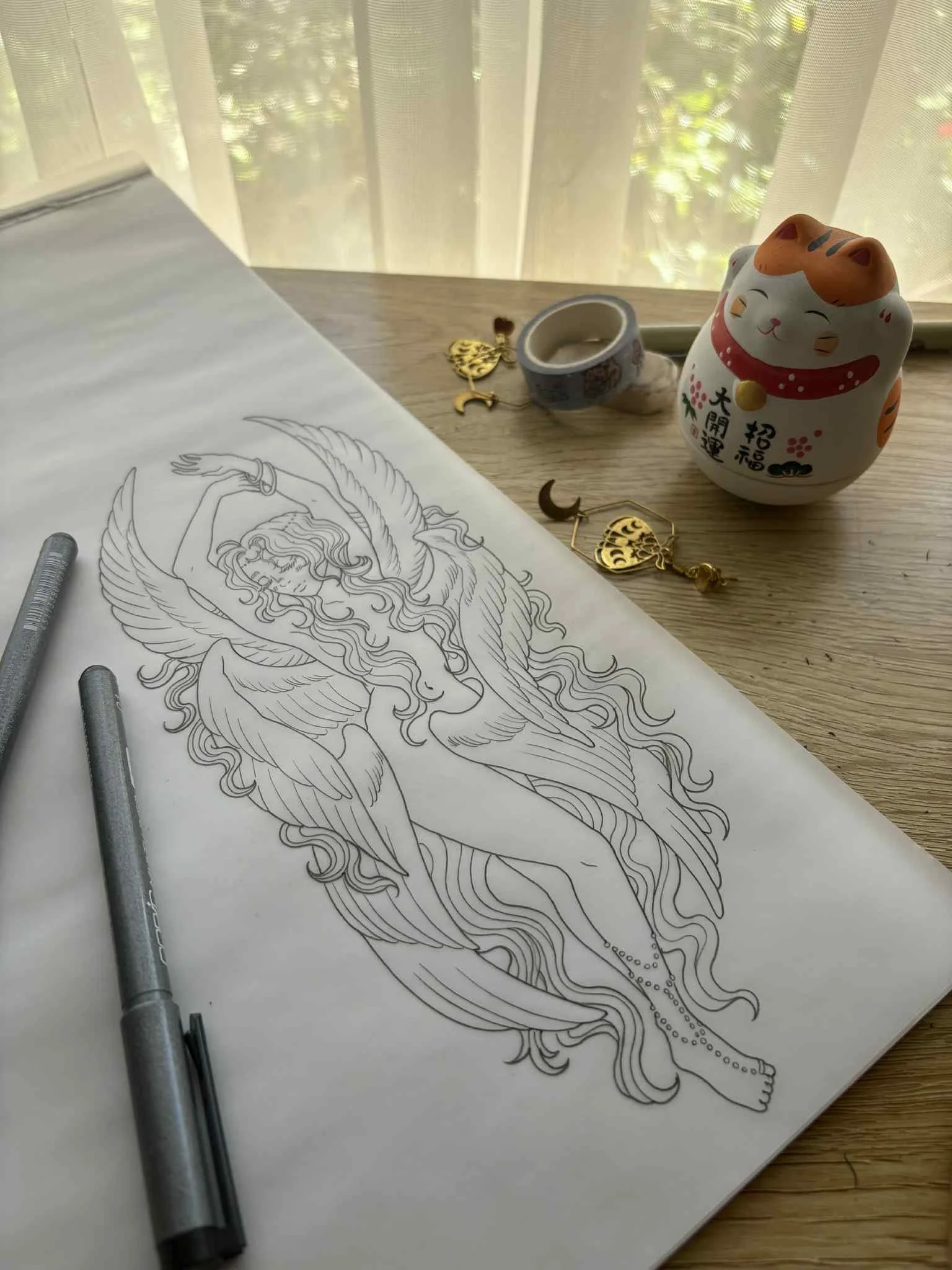 A black and white line drawing of a fairy with wings, long hair, and a serene expression, on a large sheet of paper on a wooden table. Nearby are two gray markers, a ceramic cup, a decorative keychain with a gold crescent moon and trees, a pair of earrings, a ceramic lucky cat figurine, and a washi tape dispenser. In the background, there is a window with sheer curtains and foliage outside.