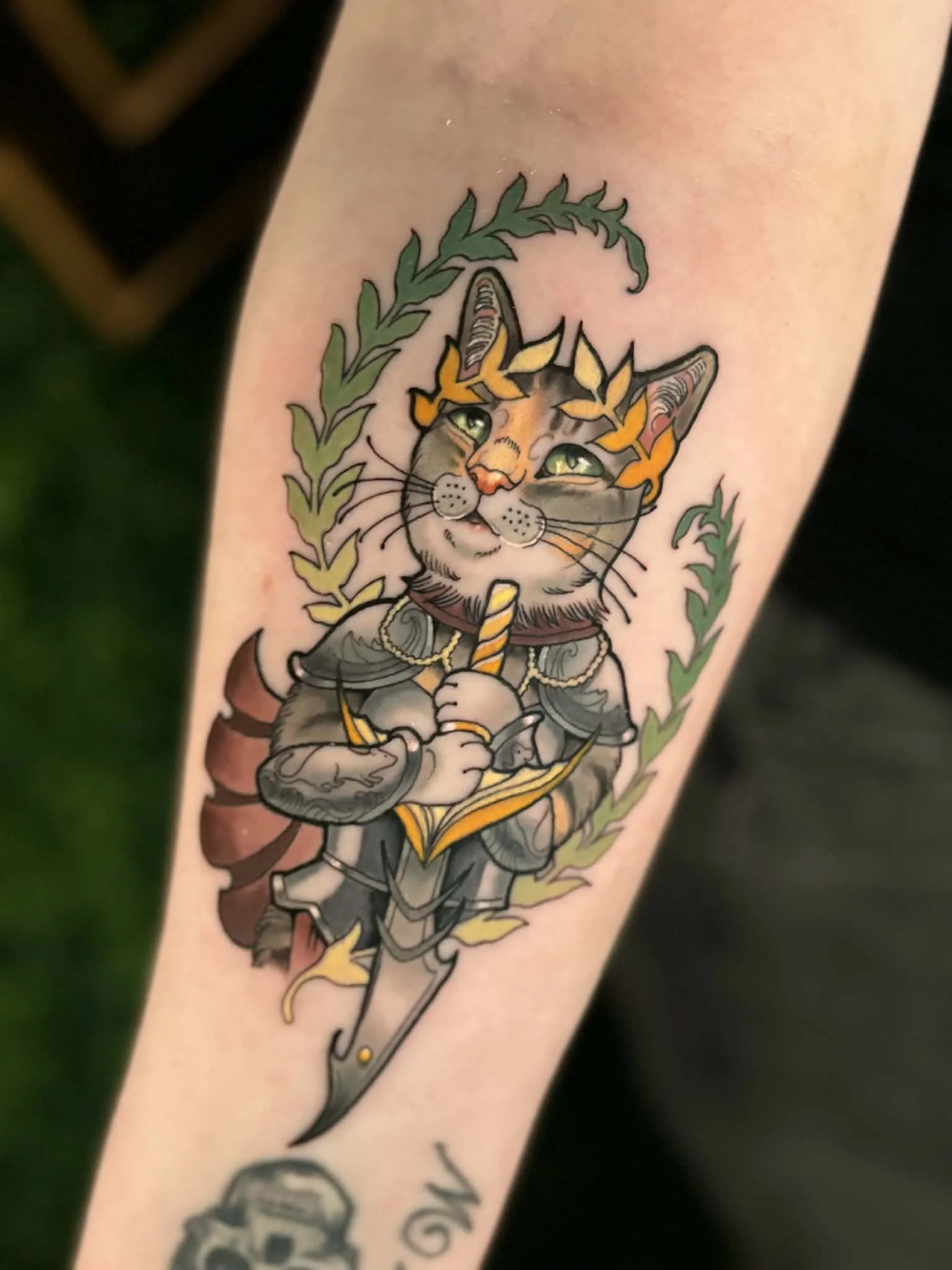 Tattoo of a cat dressed as a fantasy knight, holding a sword and a wand, with a laurel wreath surrounding its head.