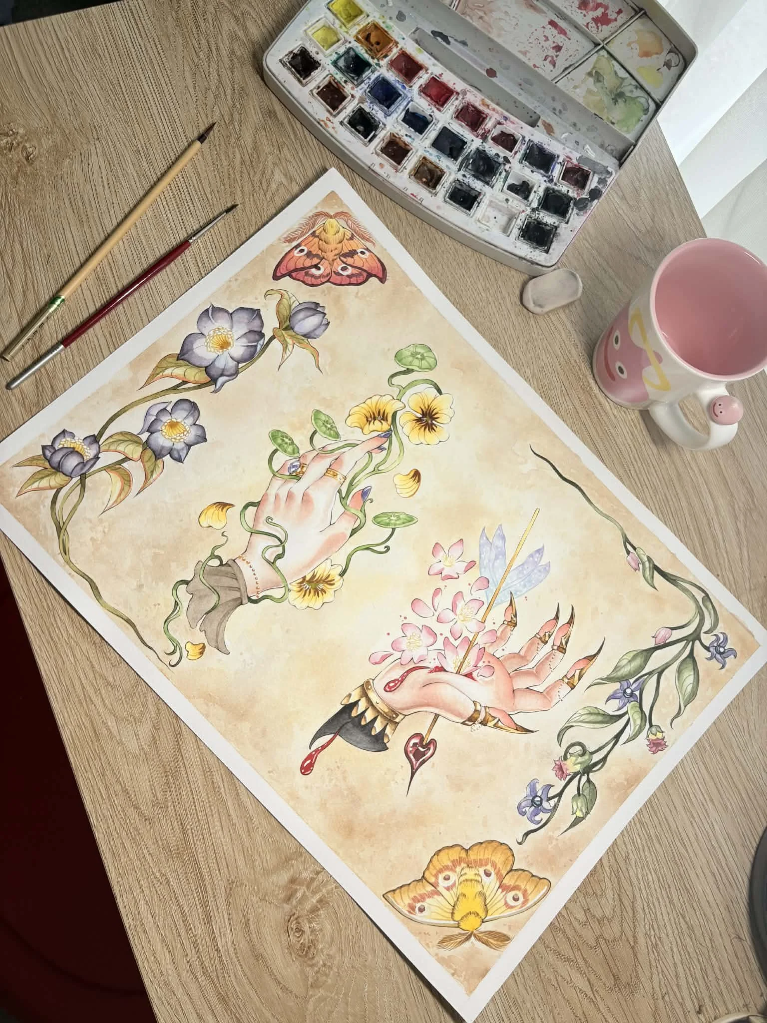 A watercolor painting on paper featuring two hands, one holding a stick with pink petals and the other reaching for yellow flowers, surrounded by vines and butterflies. The painting is on a wooden table with watercolor paints, brushes, a pink mug, and an eraser nearby.