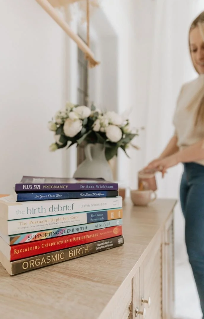 Stack of books on a wooden table, with a white vase filled with white flowers in the background. A woman is partially visible, near a bowl on the table.