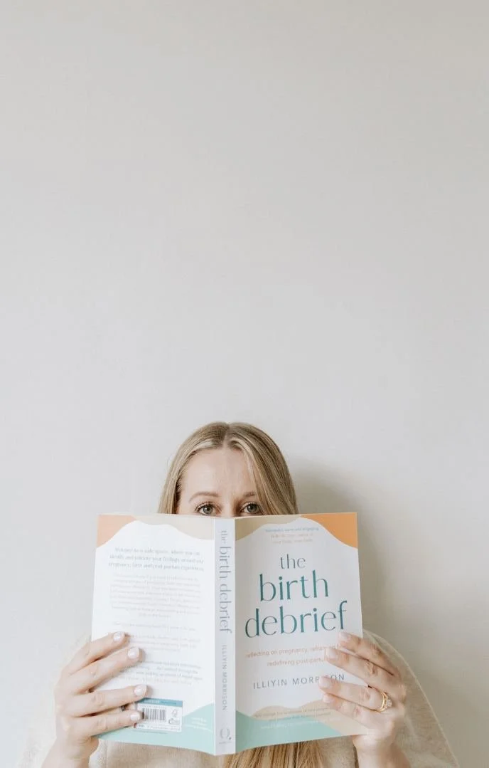 Woman holding a book titled 'The Birth Debrief' in front of her face, revealing only her eyes and forehead, against a plain light-colored wall.