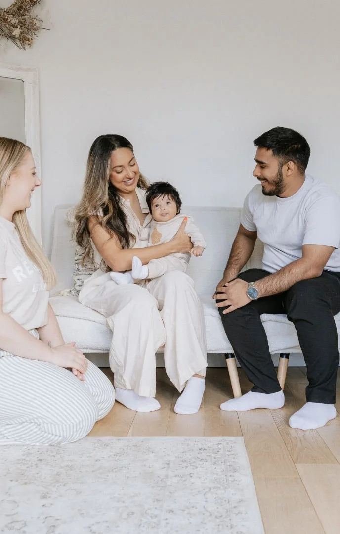 A family of four sitting on a white sofa in a living room, smiling and interacting with a young child.