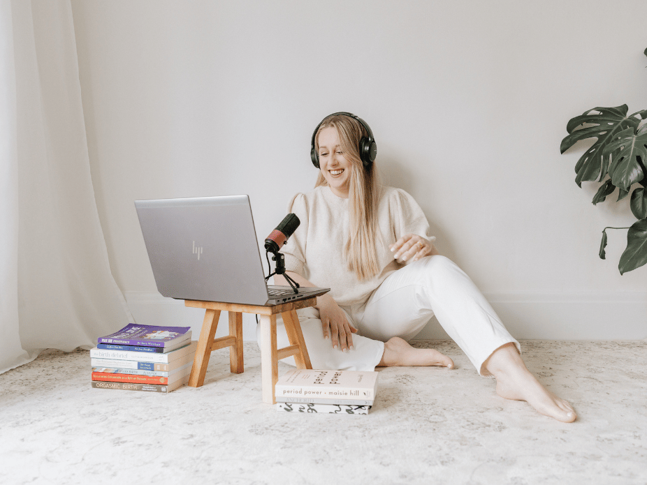 A woman with long blonde hair wearing headphones, sitting on a beige carpeted floor, smiling and recording in front of a laptop on a small wooden stool, surrounded by books, with a large green houseplant in the background.