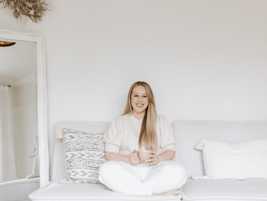 A woman sitting cross-legged on a light-colored sofa, holding a mug, smiling, in a bright, minimalistic room with white walls and decor.