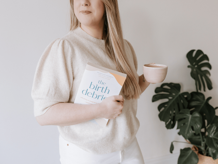 A woman holding a book titled 'the birth debrief' and a cup, standing next to a large potted Monstera plant.