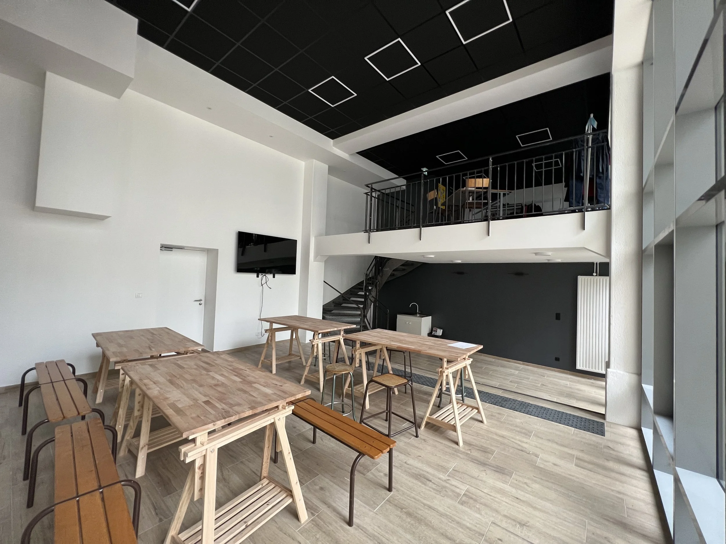 Une salle de réunion ou espace de travail moderne avec plusieurs tables en bois, des bancs en bois et en métal, un mur blanc, un mur noir en fond, une télévision murale, et un espace à l'étage supérieur avec une rampe en métal, fenêtres de grande tai