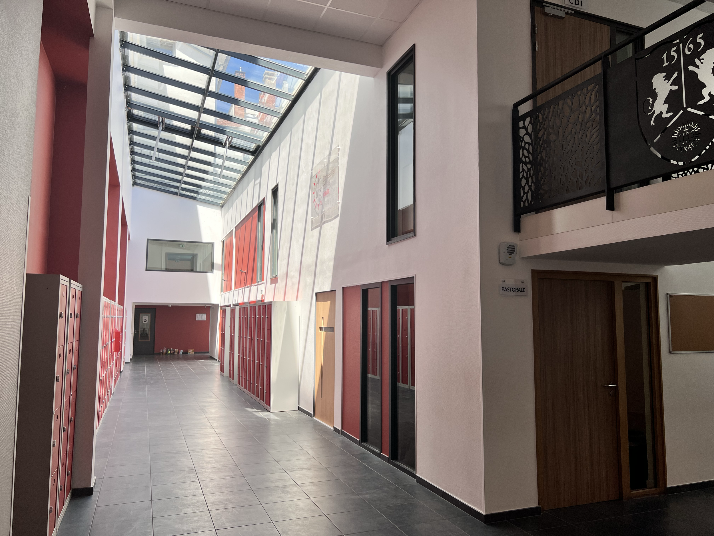 Couloir éclairé par un plafond en verre, avec des casiers rouges alignés sur les côtés et des portes en bois, dans un bâtiment moderne.