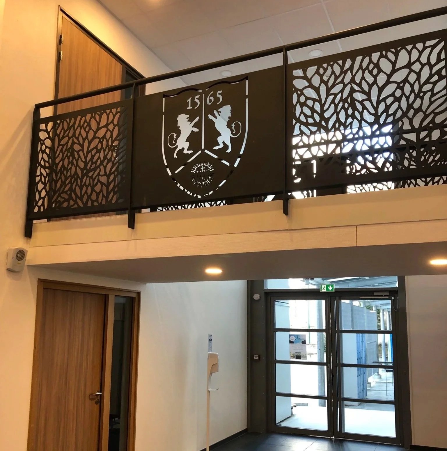 Vue intérieure d'une entrée avec porte en verre et un balcon orné d'une grille en métal noir avec un écusson historique, présentant deux lions et la date 1565.