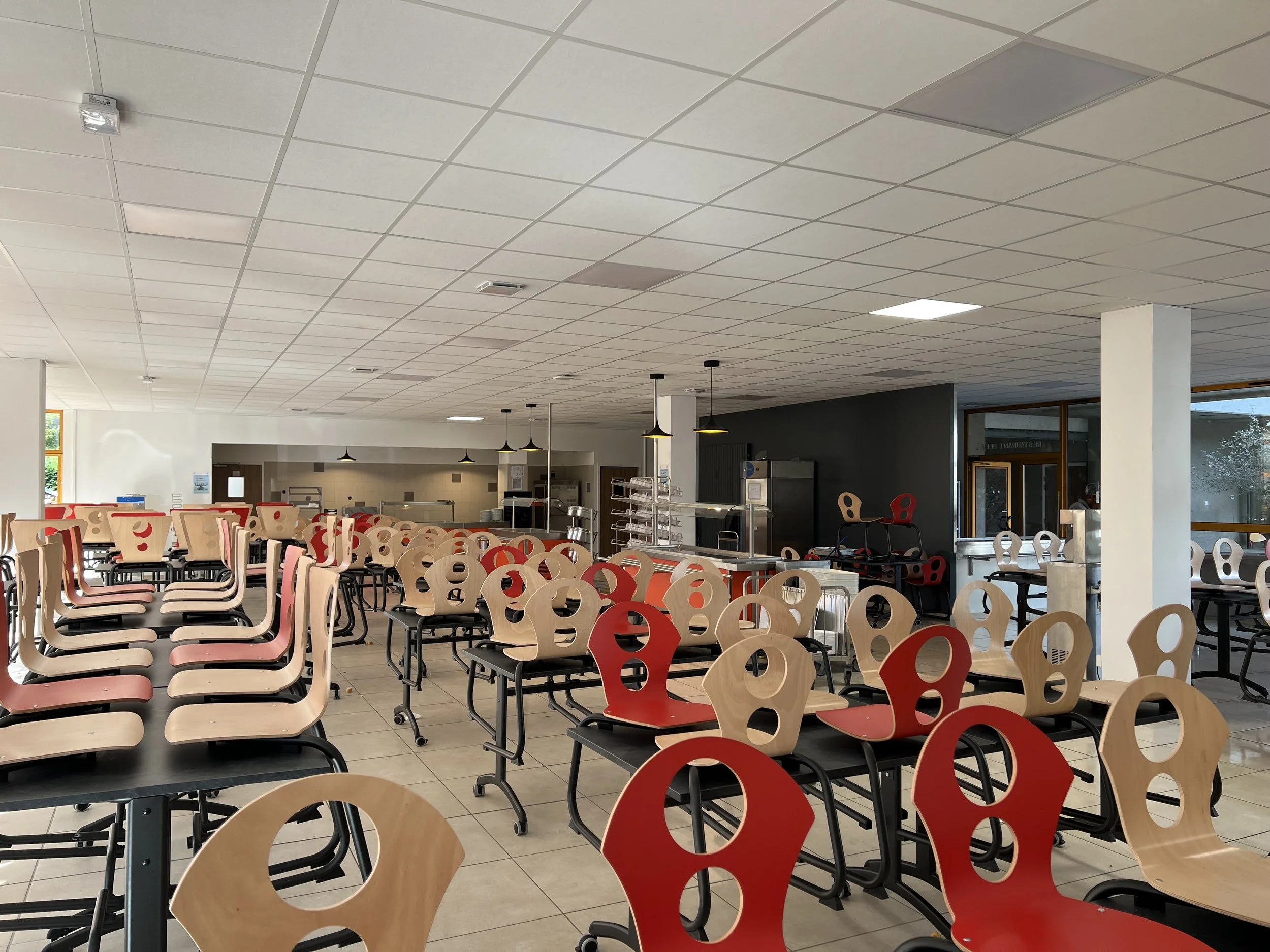 Salle à manger vide avec des chaises en bois et en plastique rouge et beiges, et des tables noires, dans un cadre lumineux avec des fenêtres et un plafond en dalles blancs.