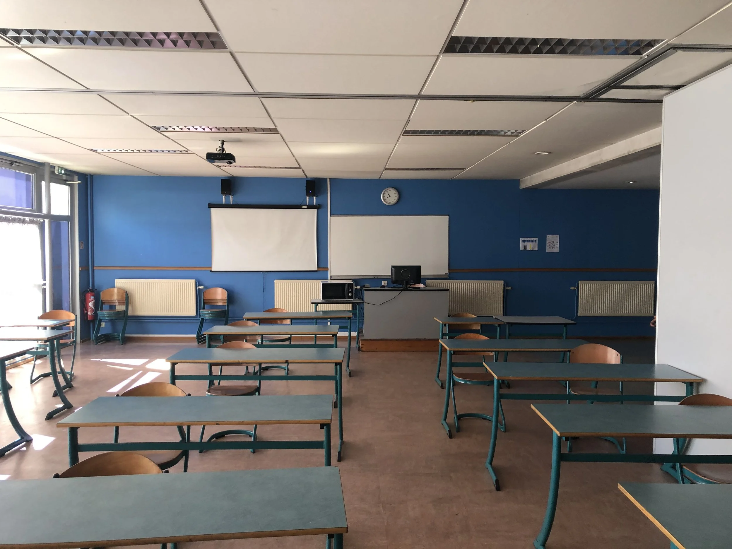 Salle de classe vide avec des bureaux, des chaises en bois, un tableau blanc, un projecteur et un écran, murs bleus, et une horloge.