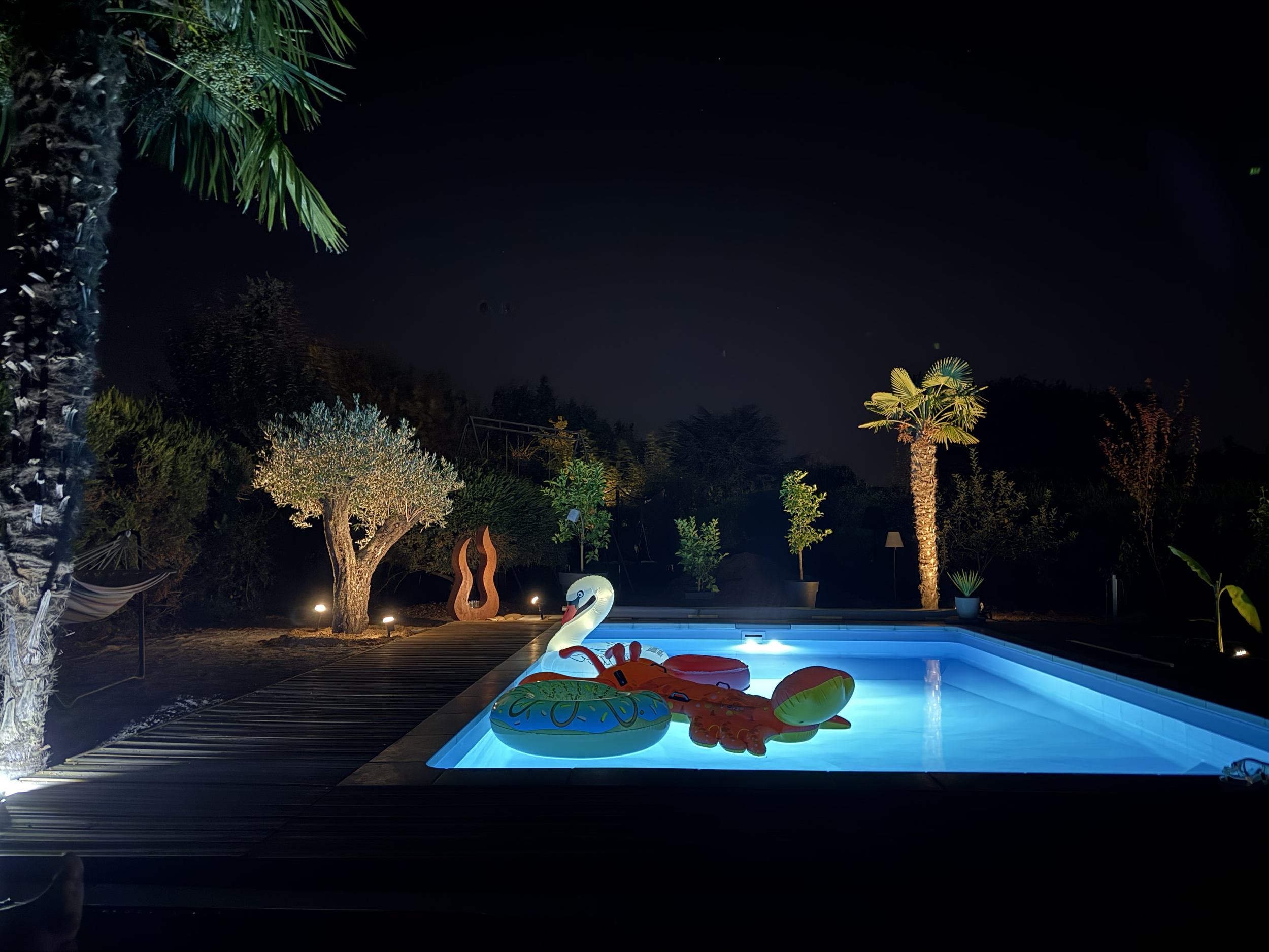 Piscine éclairée la nuit avec des jouets gonflables, palmiers, arbres, sculptures modernes et lampes autour.