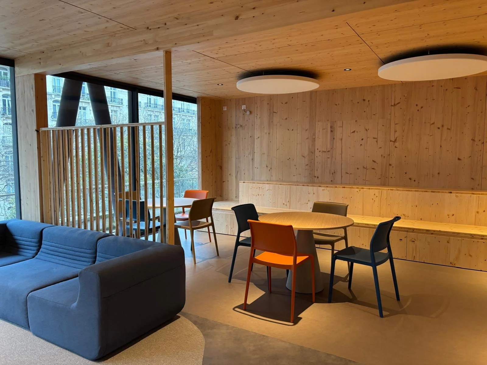 Salon moderne avec mur en bois clair, canapé gris, tables rondes en bois avec chaises colorées, et fenêtres grandes laissant entrer la lumière naturelle.