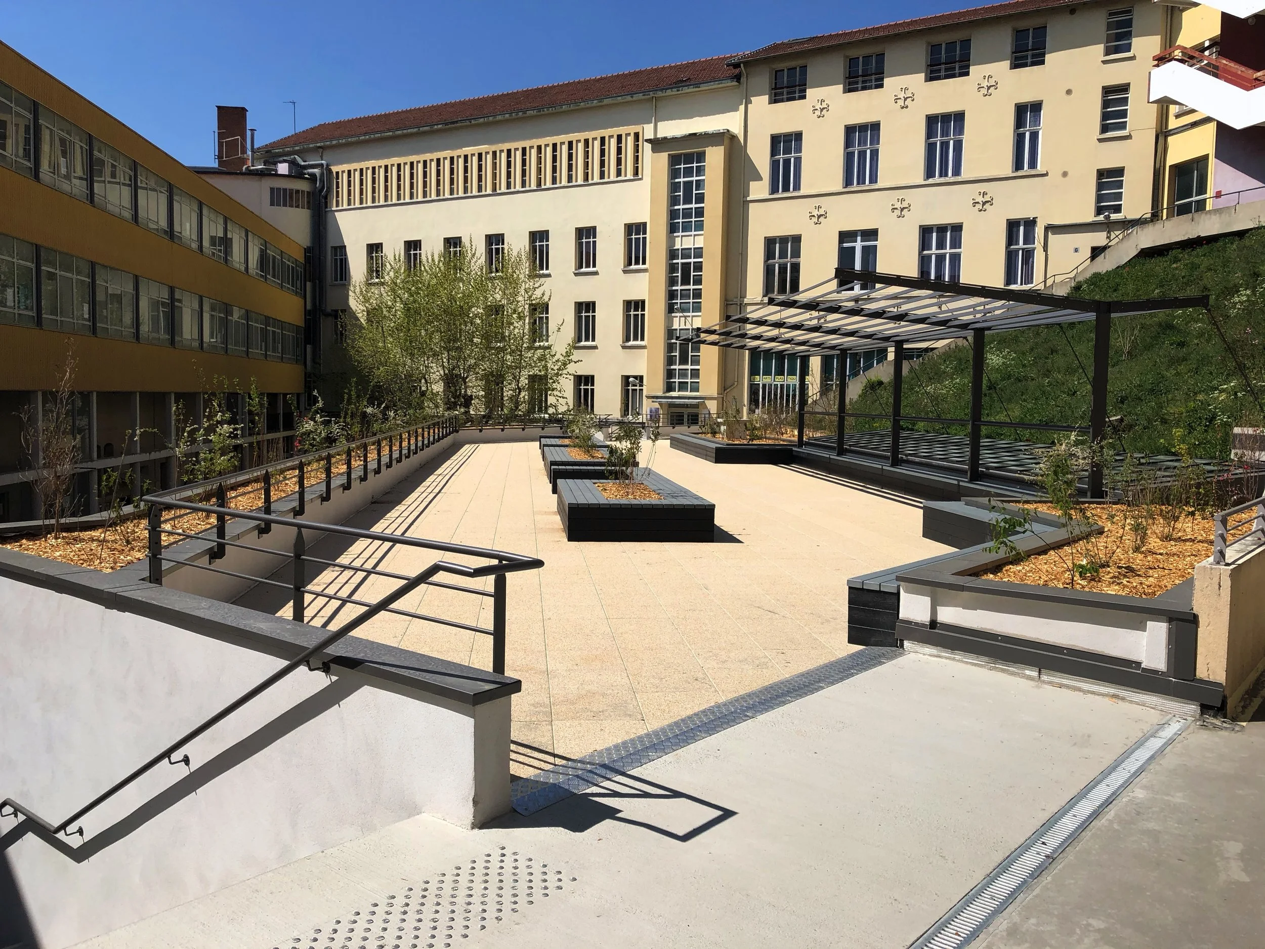 Cour extérieure d'un bâtiment avec des allées pavées, des jardinières et une structure en métal pour se reposer ou se protéger du soleil. Bâtiments en arrière-plan avec plusieurs fenêtres et une coloration beige et jaune.