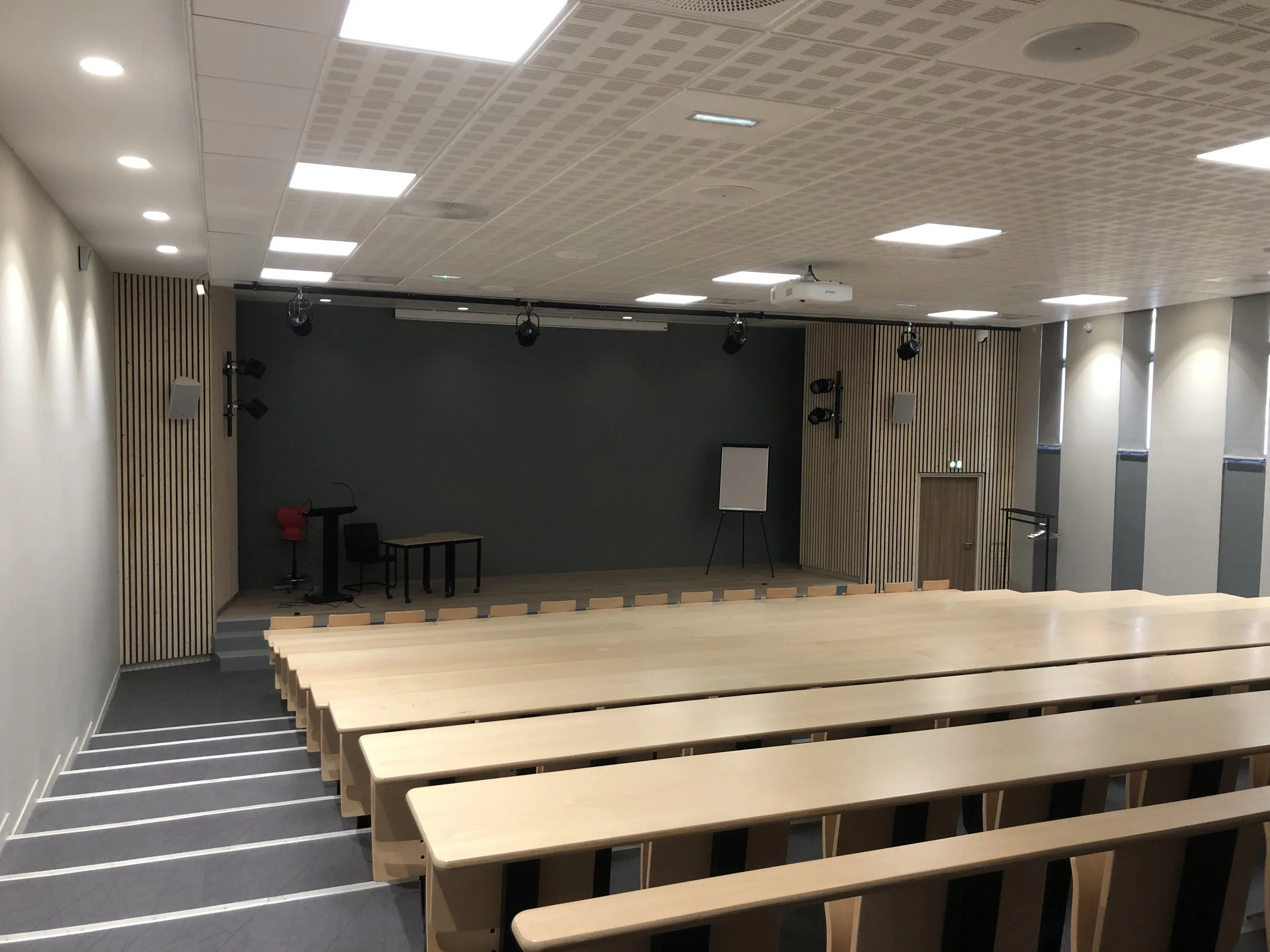 Une salle de conférence ou d'auditorium vide avec des rangées de bancs en bois, un espace de scène avec un tableau blanc et du matériel de sonorisation, murs en bois et plafond avec éclairage encastré.