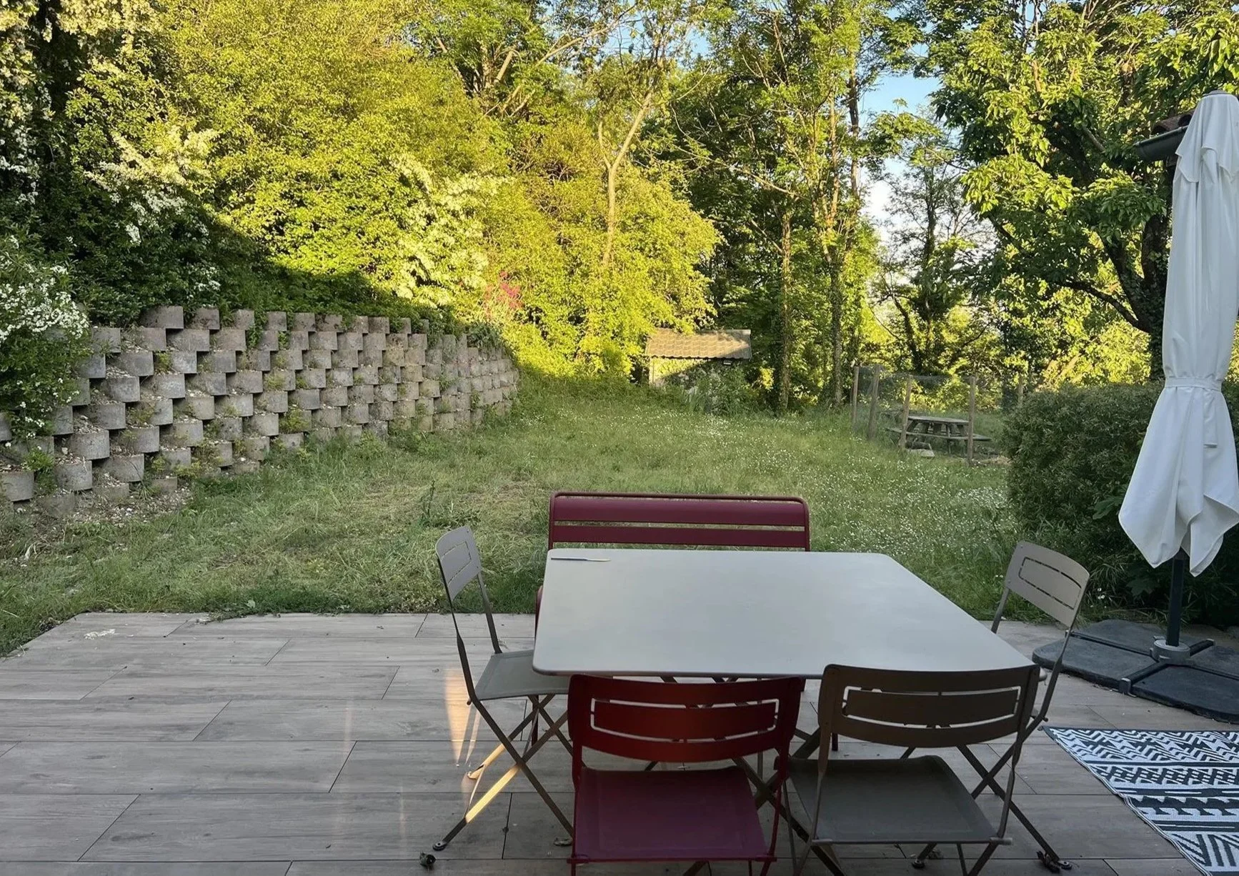 Terrasse avec table, chaises, parasol fermé, jardin verdoyant avec arbres et tonnelle en arrière-plan.