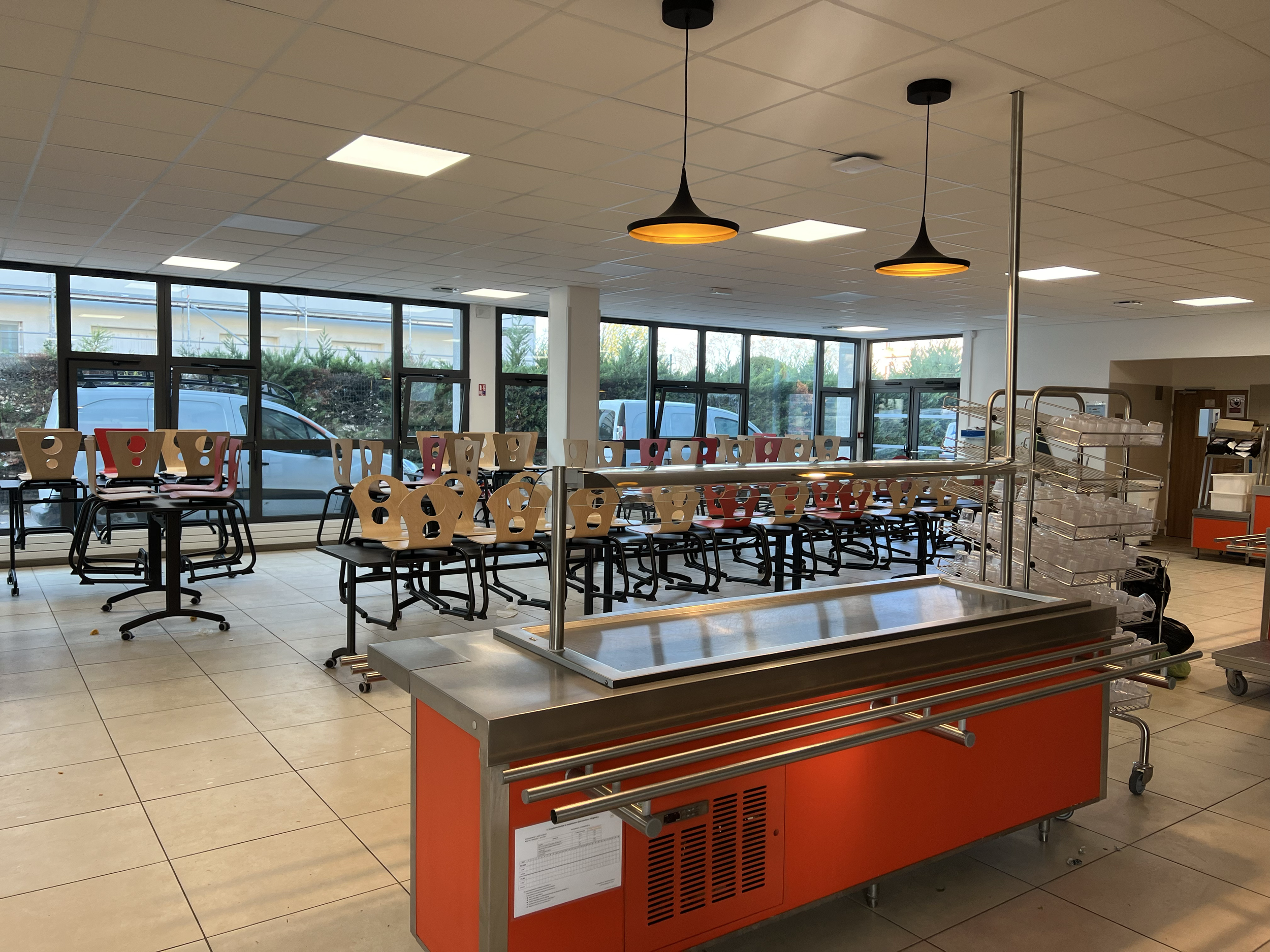 Salle de restaurant ou de cafétéria vide avec des chaises empilées sur des tables, un meuble de service en acier inoxydable orange, de grandes fenêtres avec des voitures garées à l'extérieur, et un plafond avec des luminaires suspendus