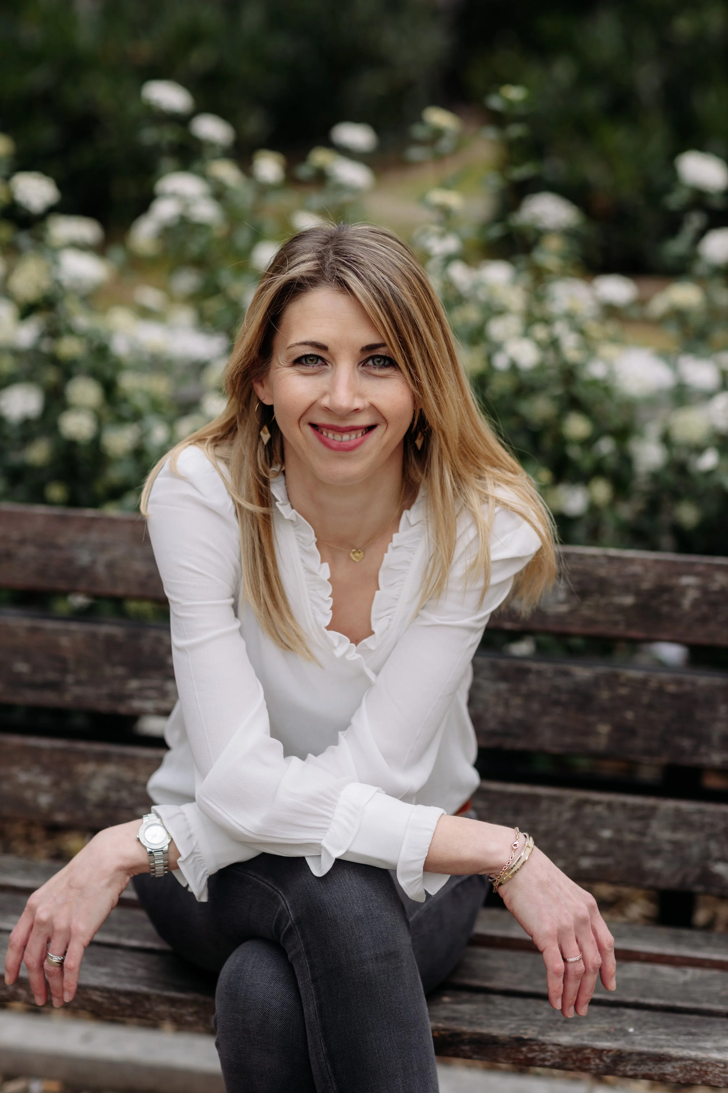 Une femme souriante, assise sur un banc en bois dans un jardin avec des buissons de fleurs blanches en arrière-plan.