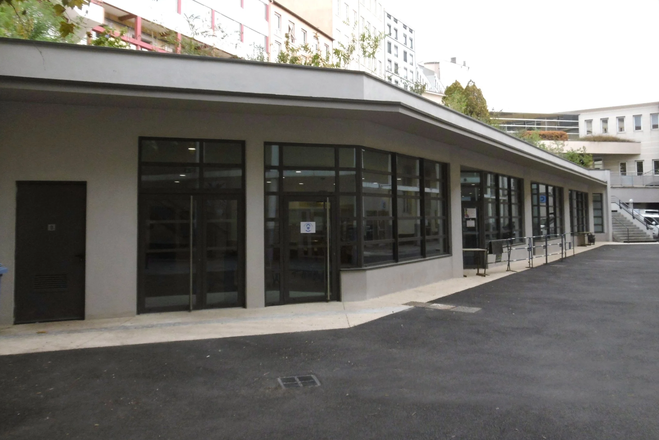 Facilité sportive ou centre communautaire avec de grandes fenêtres en verre et un accès piétonnier, en milieu urbain avec des immeubles résidentiels en arrière-plan.