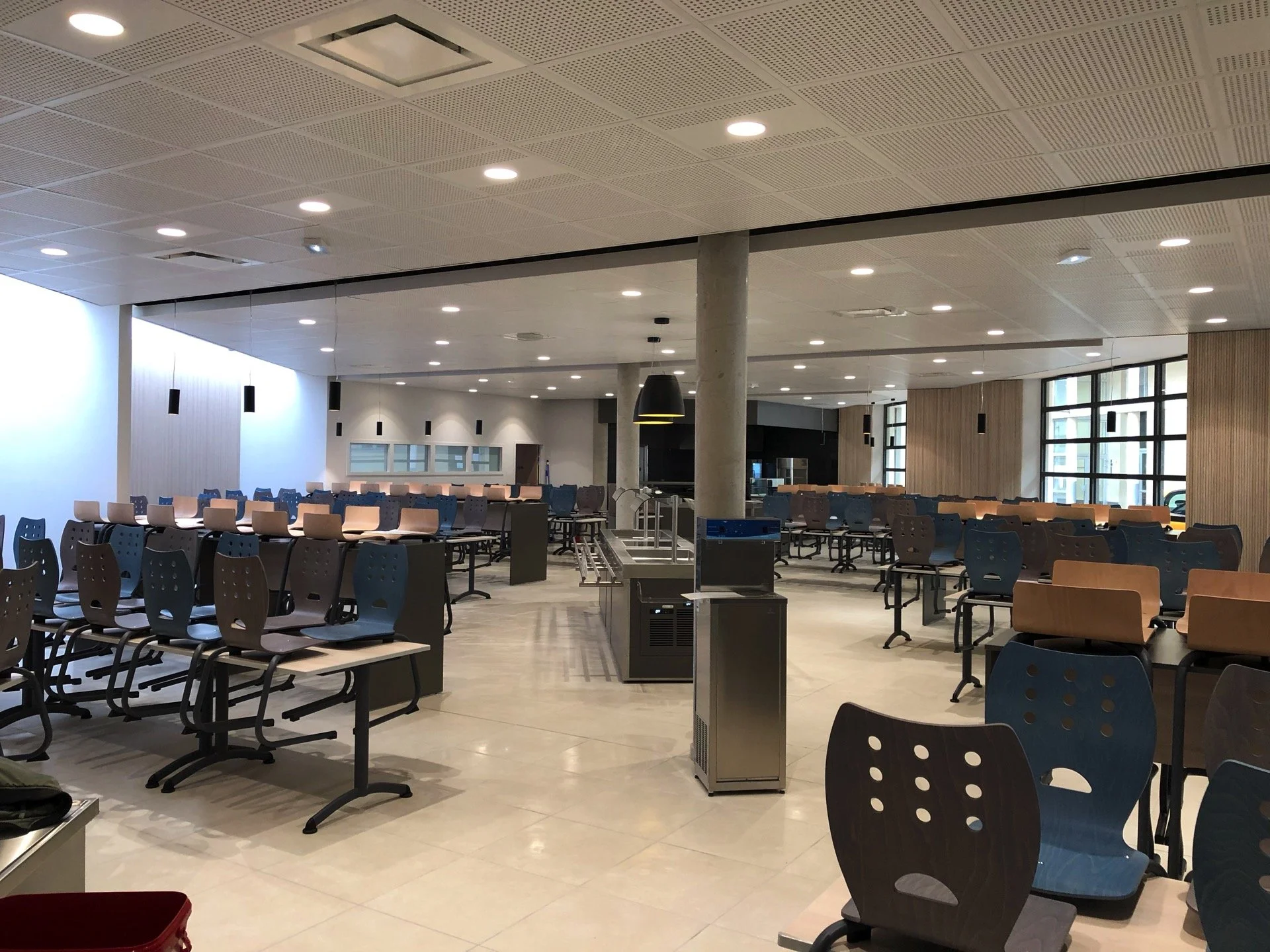 Salle de restauration ou cafétéria avec plusieurs tables et chaises en bois et en plastique, lumière suspendue, fenêtres larges laissant entrer la lumière naturelle, mobilier moderne.
