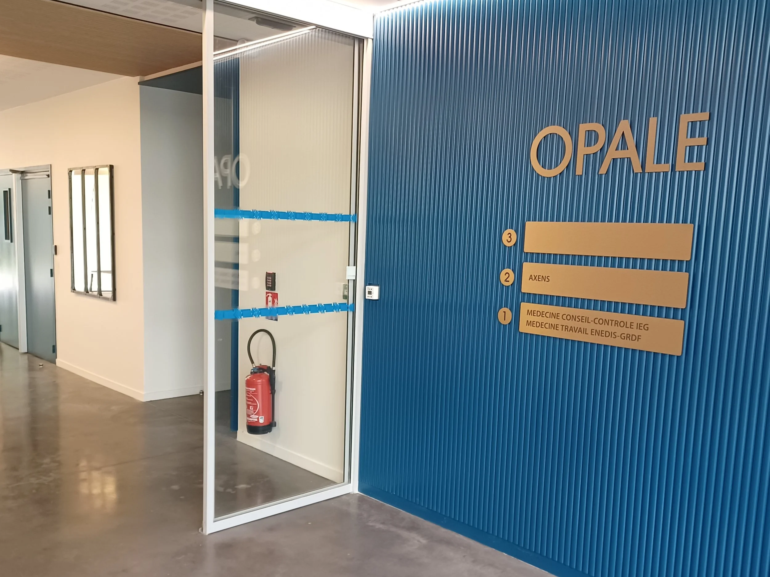 Mur bleu avec un panneau indiquant les noms et numéros des bureaux, et une porte vitrée avec un extincteur à l'intérieur.