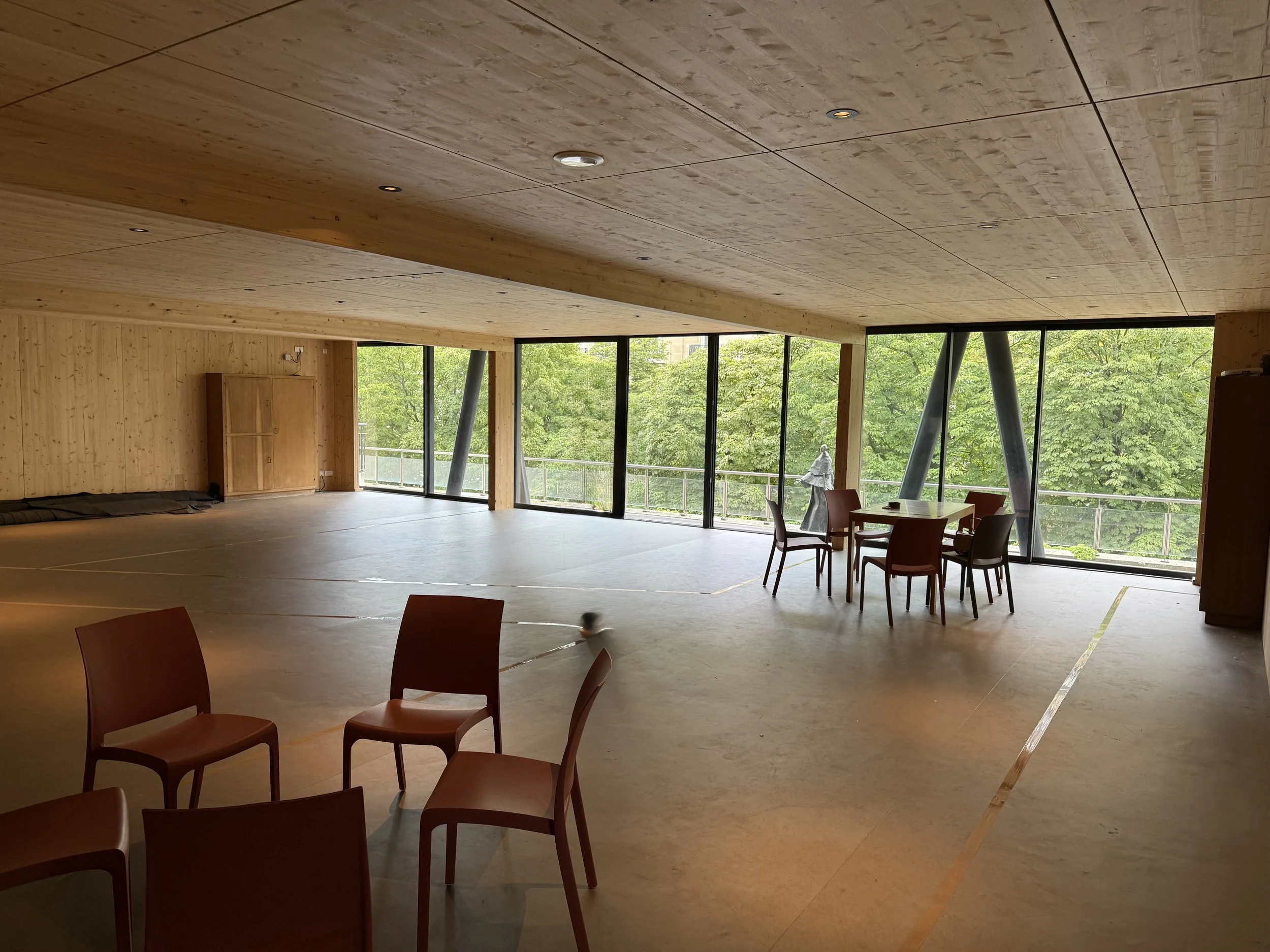 Salle spacieuse avec plafond en bois, grande baie vitrée donnant sur des arbres verts, chaises en bois et une table à manger.