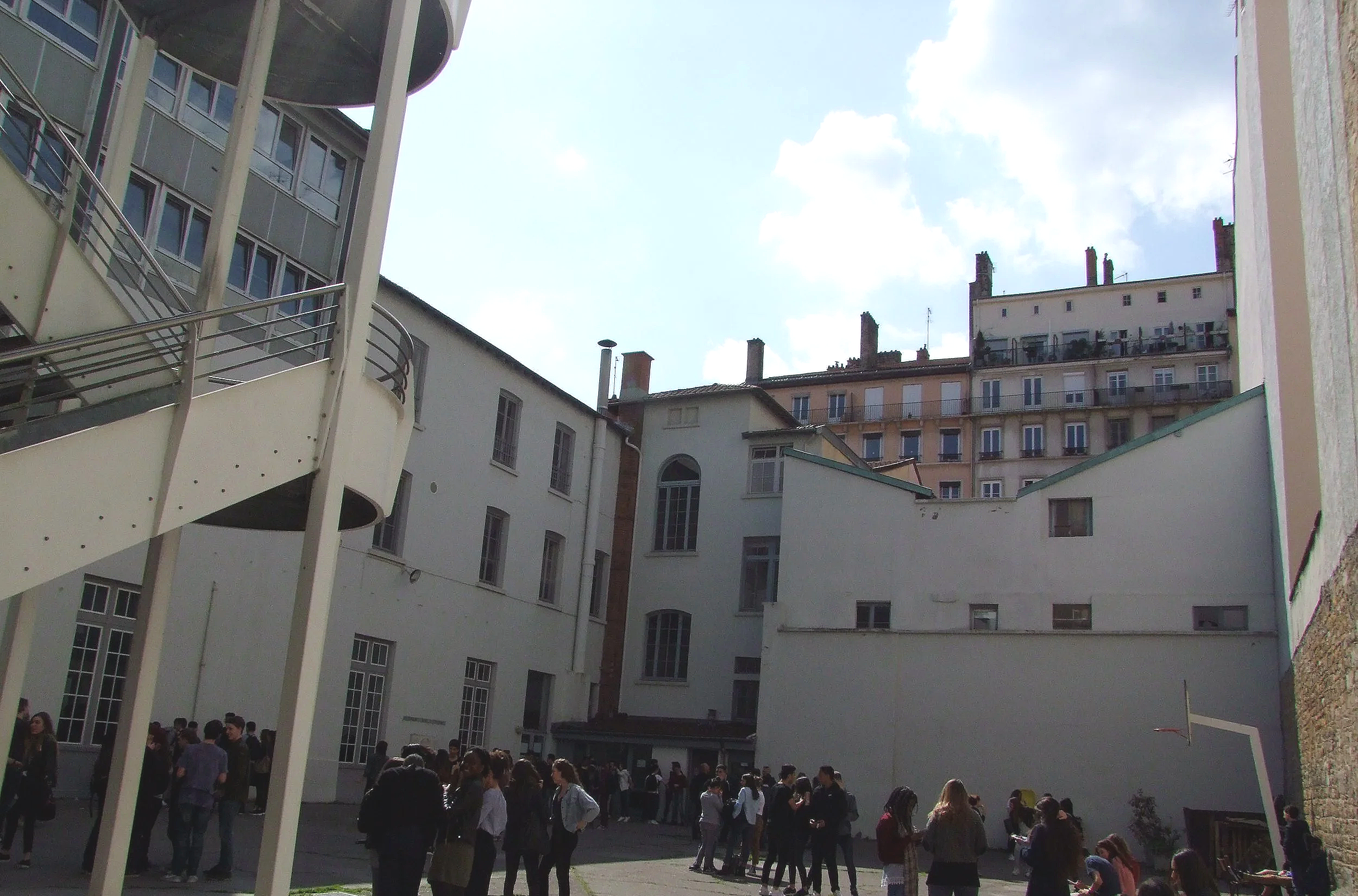 Une cour intérieure avec des étudiants rassemblés, entourée de bâtiments blancs avec plusieurs fenêtres, sous un ciel partiellement nuageux.