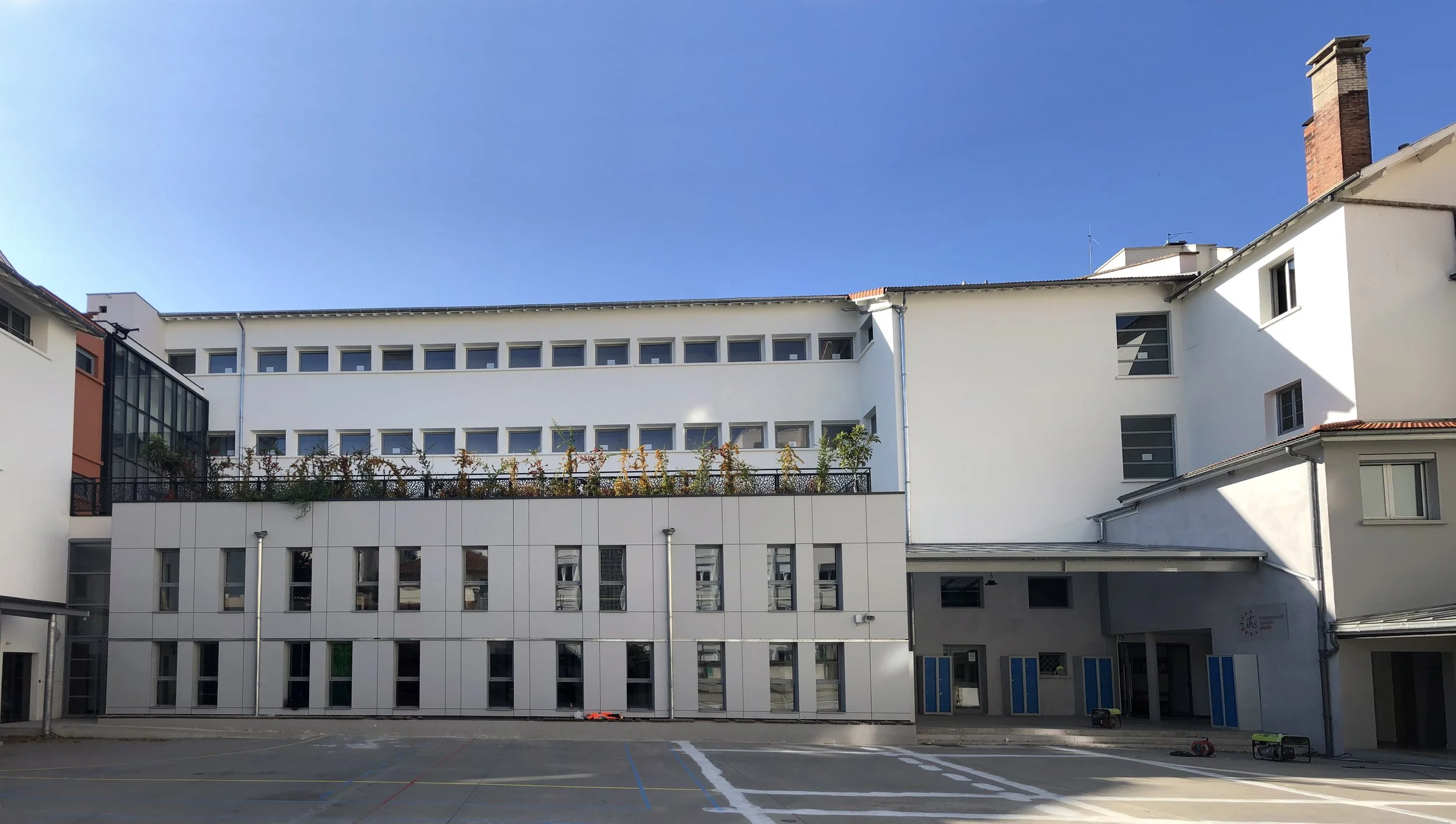 Vue d'une cour d'école ou d'un bâtiment éducatif moderne avec plusieurs étages, fenêtre et un toit en pente, sous un ciel clair et bleu.