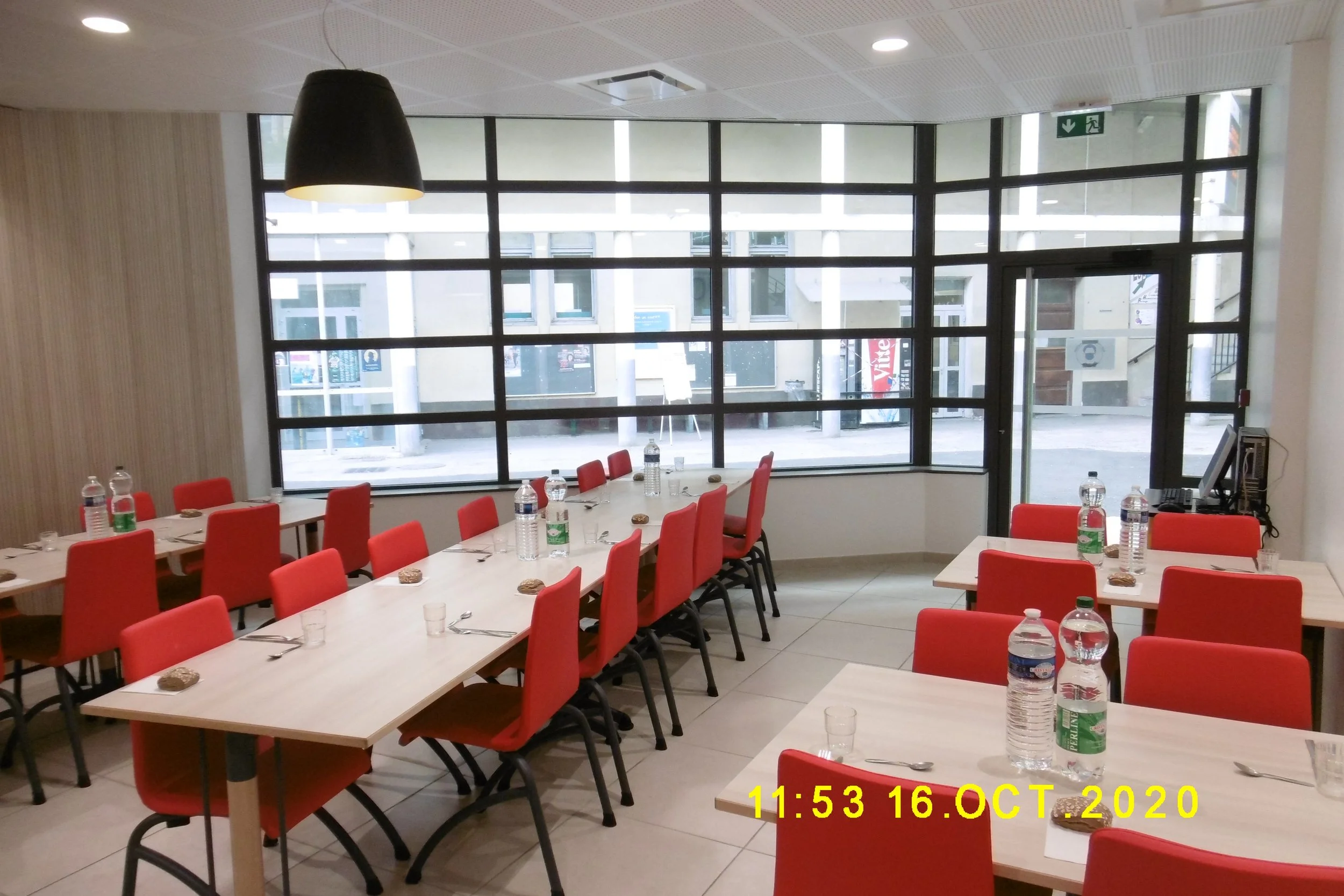 Salle de réunion avec tables et chaises rouges, bouteilles d'eau, verres et assiettes, grande fenêtre en arrière-plan, horodatage 16 octobre 2020, 11h53.