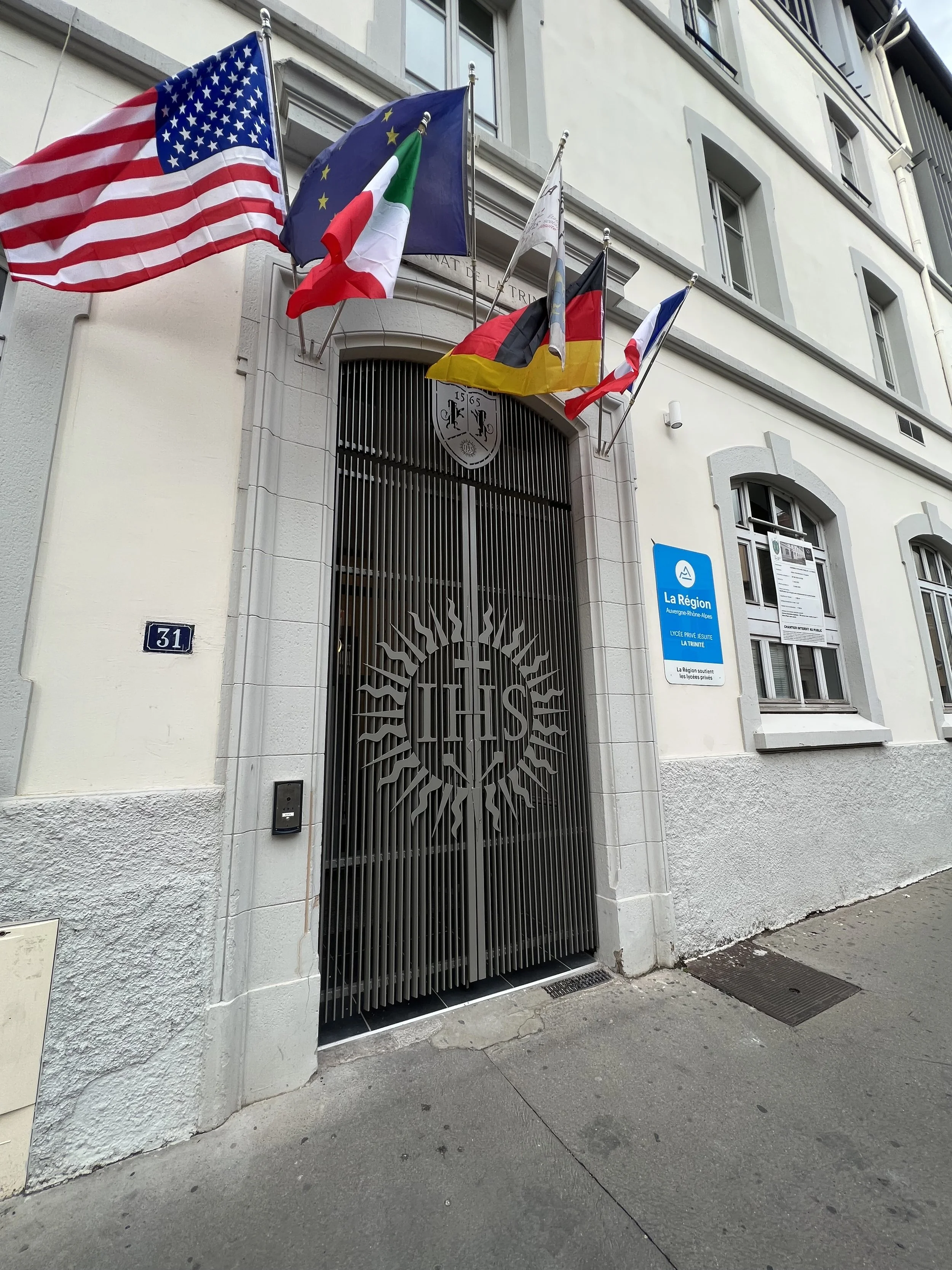 Façade d'un bâtiment avec plusieurs drapeaux internationaux suspendus au-dessus de l'entrée, y compris ceux des États-Unis, de l'Union Européenne, de l'Italie, de l'Allemagne, de la France, et d'une autre nation. La porte d'entrée est en métal avec u