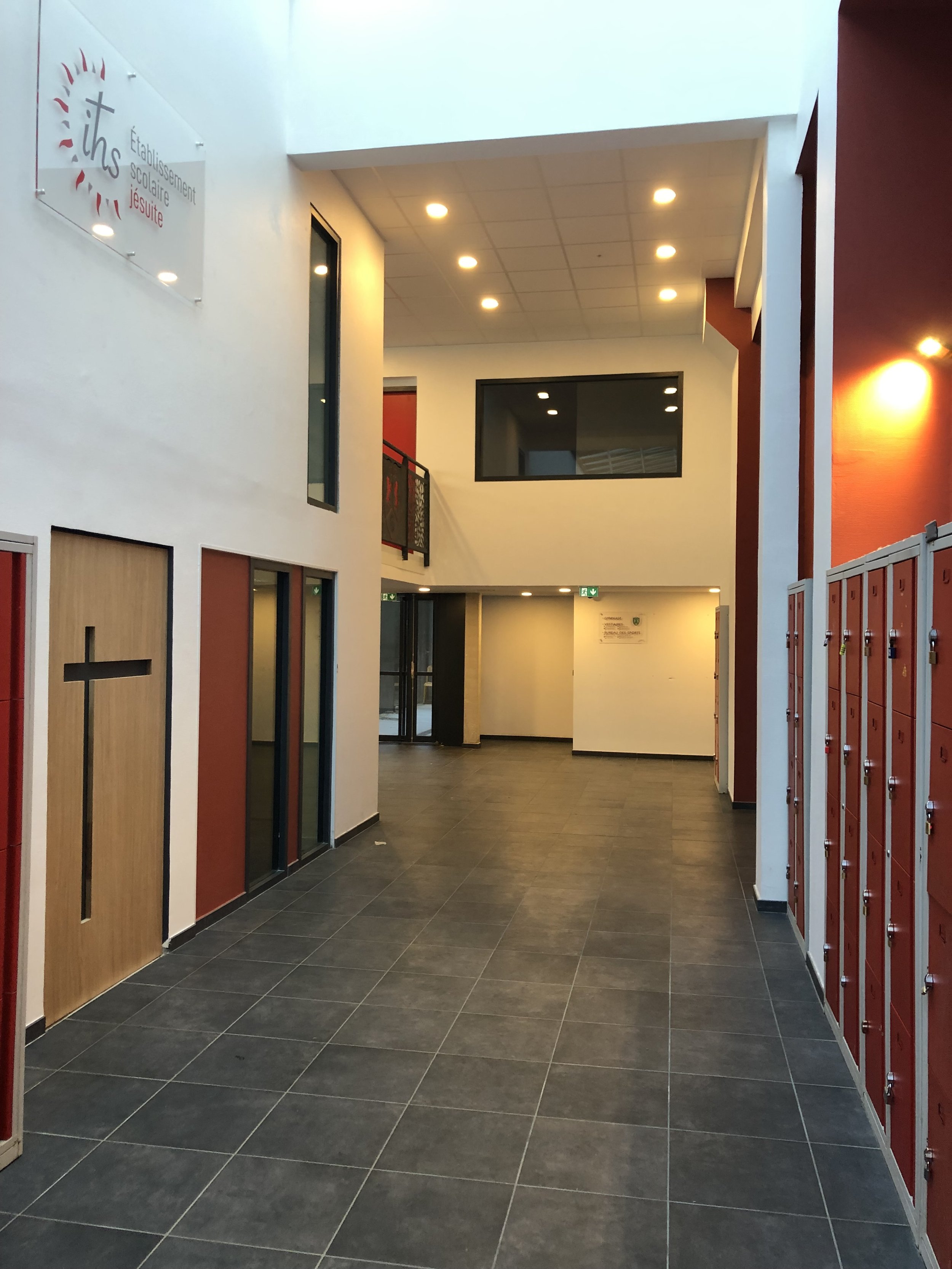 Intérieur d'un établissement scolaire avec des casiers rouges à droite, un panneau avec une croix à gauche, et l'inscription 'Établissement scolaire Jésuite' sur un mur à gauche.