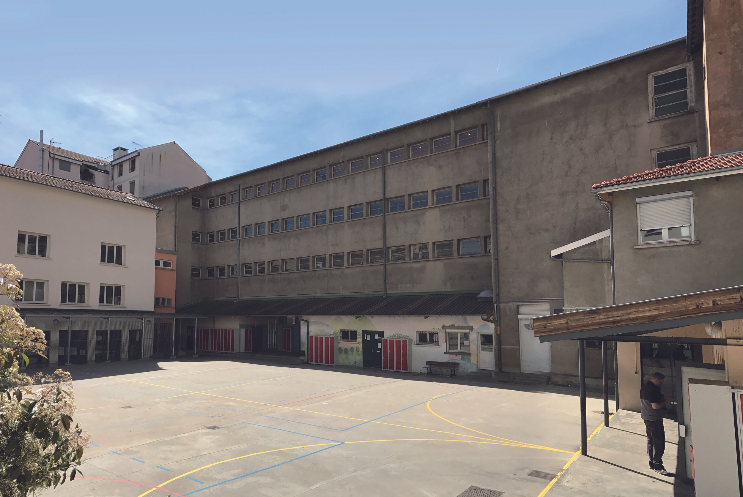 Une cour d'école entourée d'immeubles résidentiels, avec un sol en béton marqué par des lignes de couleur pour le sport, et une personne près d'un bâtiment à droite.