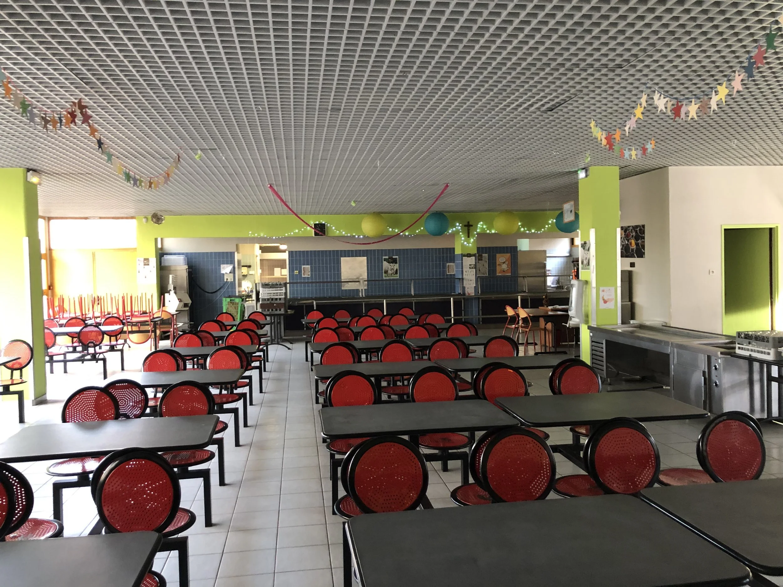 Salle de cantine vide avec tables noires et chaises rouges, décorations colorées suspendues au plafond, coin restauration avec évier et équipements en arrière-plan, fenêtre laissant entrer la lumière naturelle.