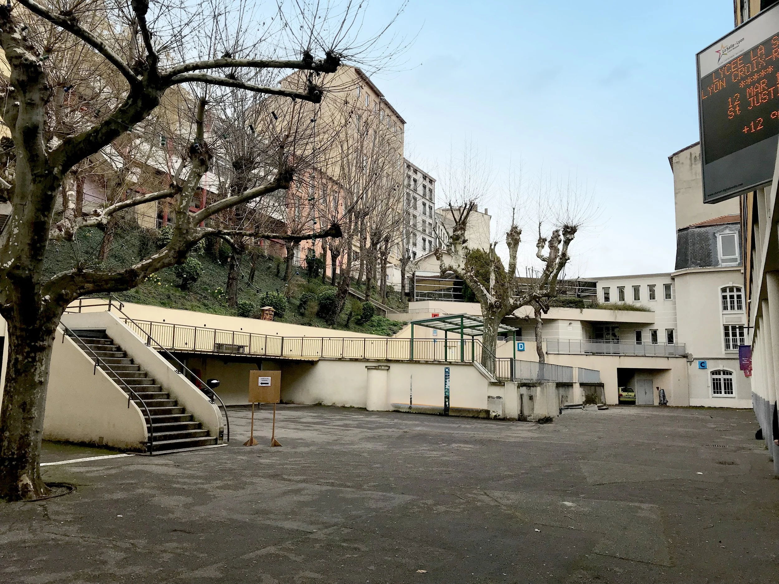 Un parking en plein air avec quelques arbres sans feuilles, des bâtiments en arrière-plan, et un panneau d'information numérique à droite.