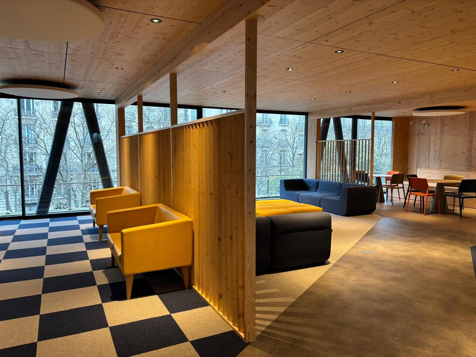 Un intérieur moderne avec des murs en bois, des fenêtres en verre laissant entrer la lumière naturelle, et des meubles de salon colorés dont des fauteuils jaunes, un canapé bleu, un canapé noir, et une table avec des chaises colorées.