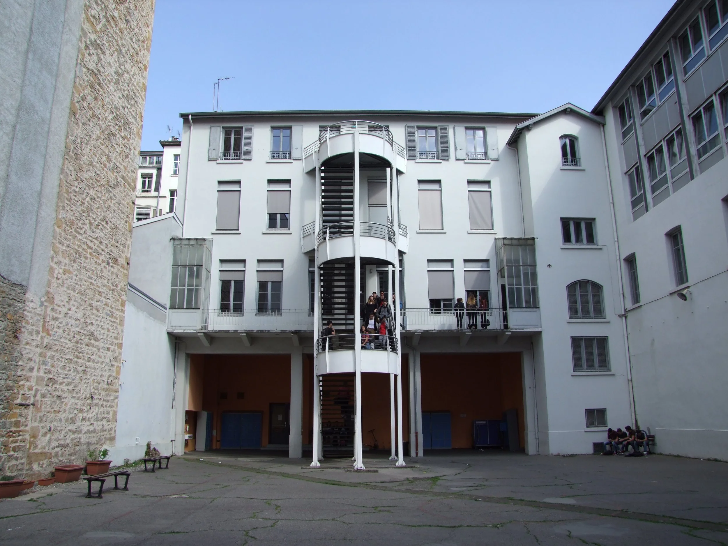 Cour intérieure d'un bâtiment résidentiel blanc avec un escalier en colimaçon central, plusieurs fenêtres, et des personnes assises ou debout.
