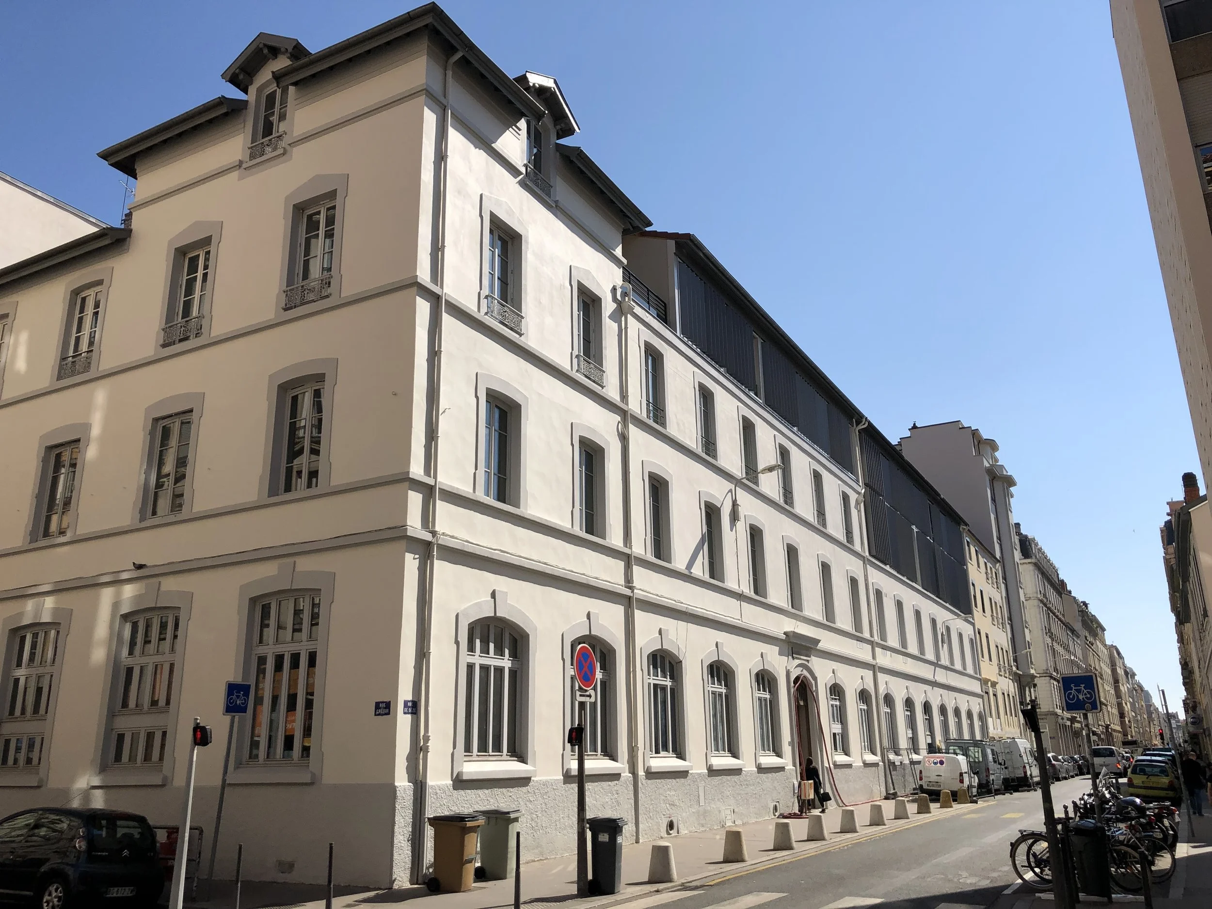 Bâtiment blanc à plusieurs étages avec fenêtres, situé dans une rue urbaine sous un ciel bleu clair.