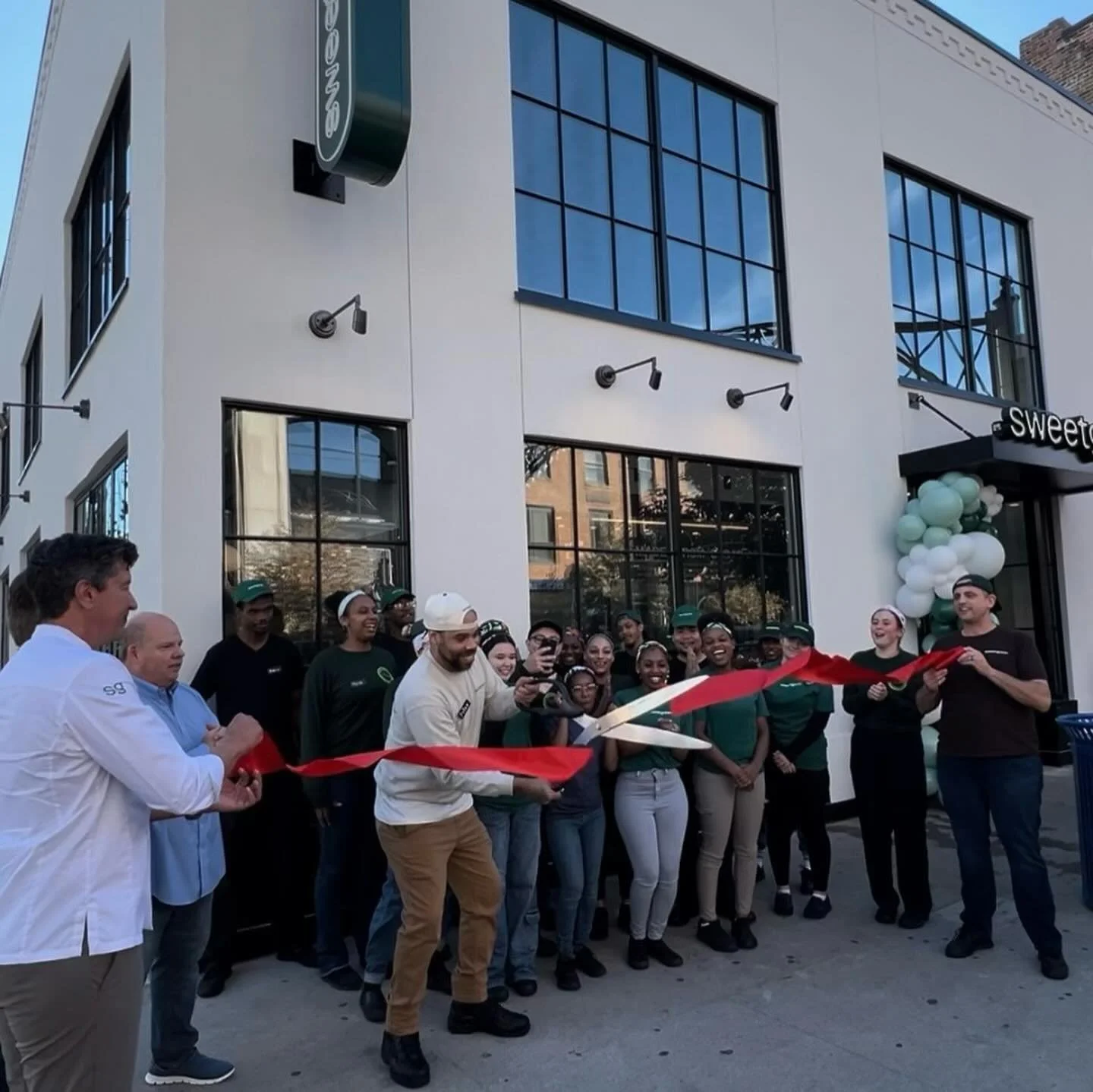 Cérémonie inauguration franchise de coupe de ruban devant un bâtiment avec un groupe de personnes souriantes, incluant des employés et un chef, en train de célébrer l'ouverture d'un nouveau commerce, décorations de ballons blancs et verts.