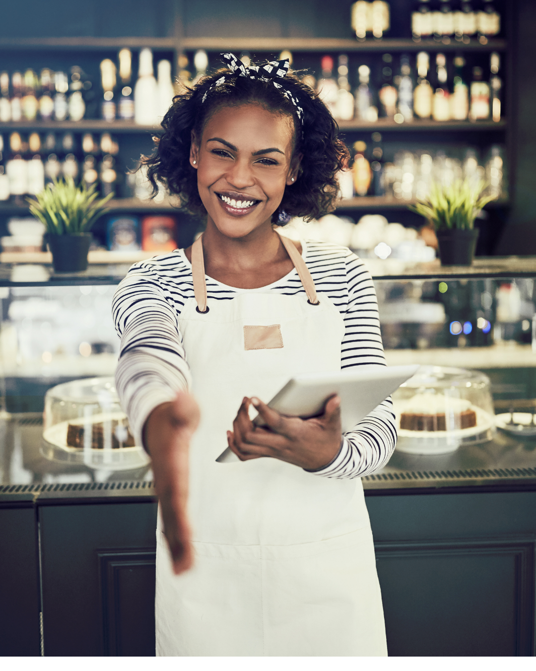 Femme serveuse dans un café, souriant et tendant la main, tenant une tablette, arrière-plan avec des étagères remplies de bouteilles et des pâtisseries dans un présentoir.