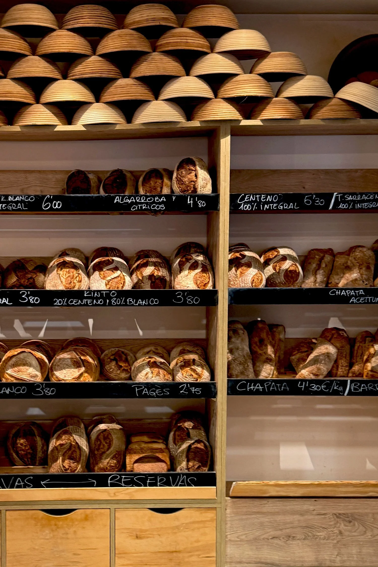 Estantes con diferentes tipos de pan artesanal y etiquetas con nombres y precios en un establecimiento de panadería.